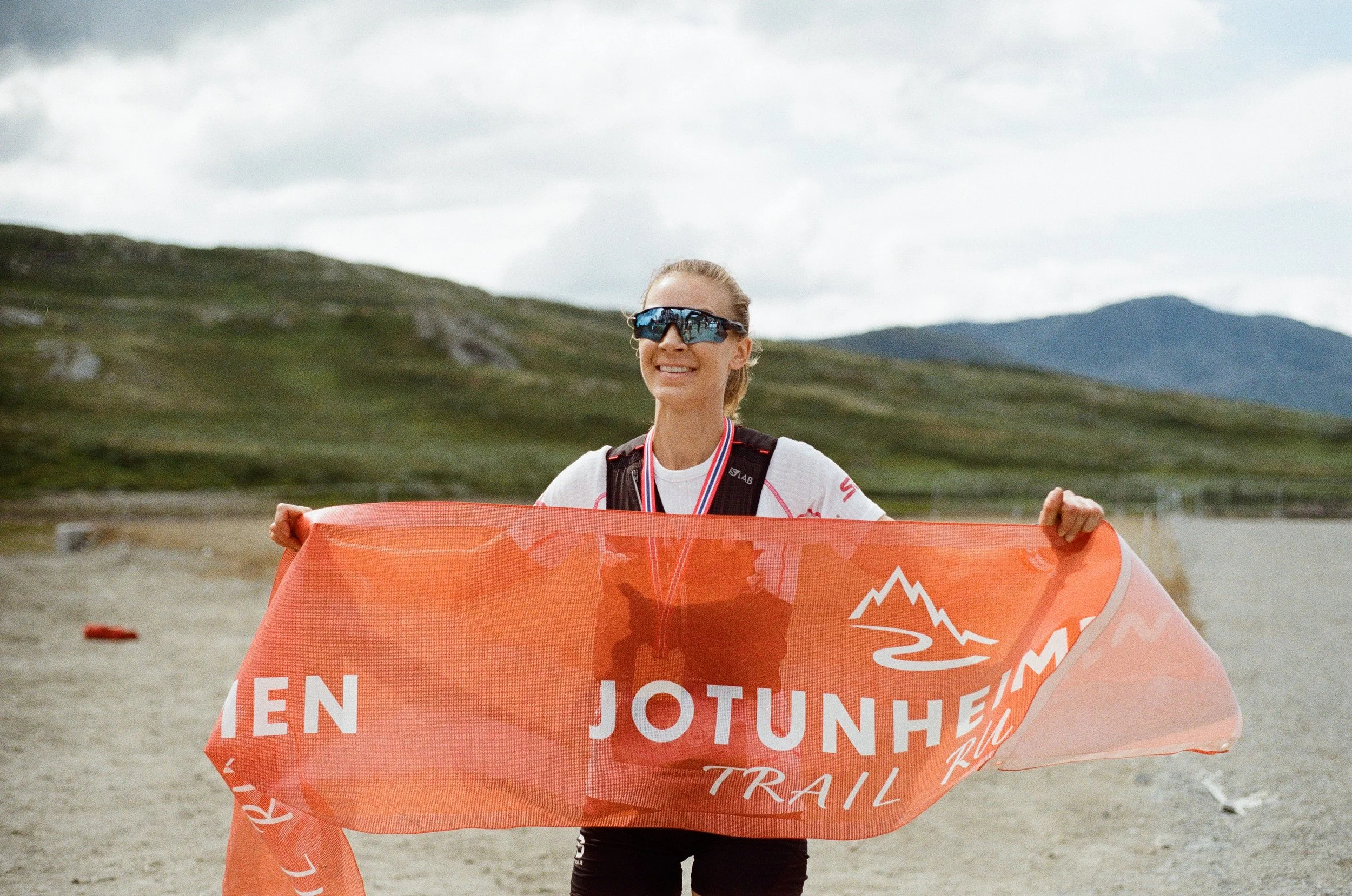 Female trail runner holding a Jotunheimen Trail Run banner, wearing sunglasses, a medal around her neck, and a hydration pack, on a mountain trail with hills and cloudy sky in the background.