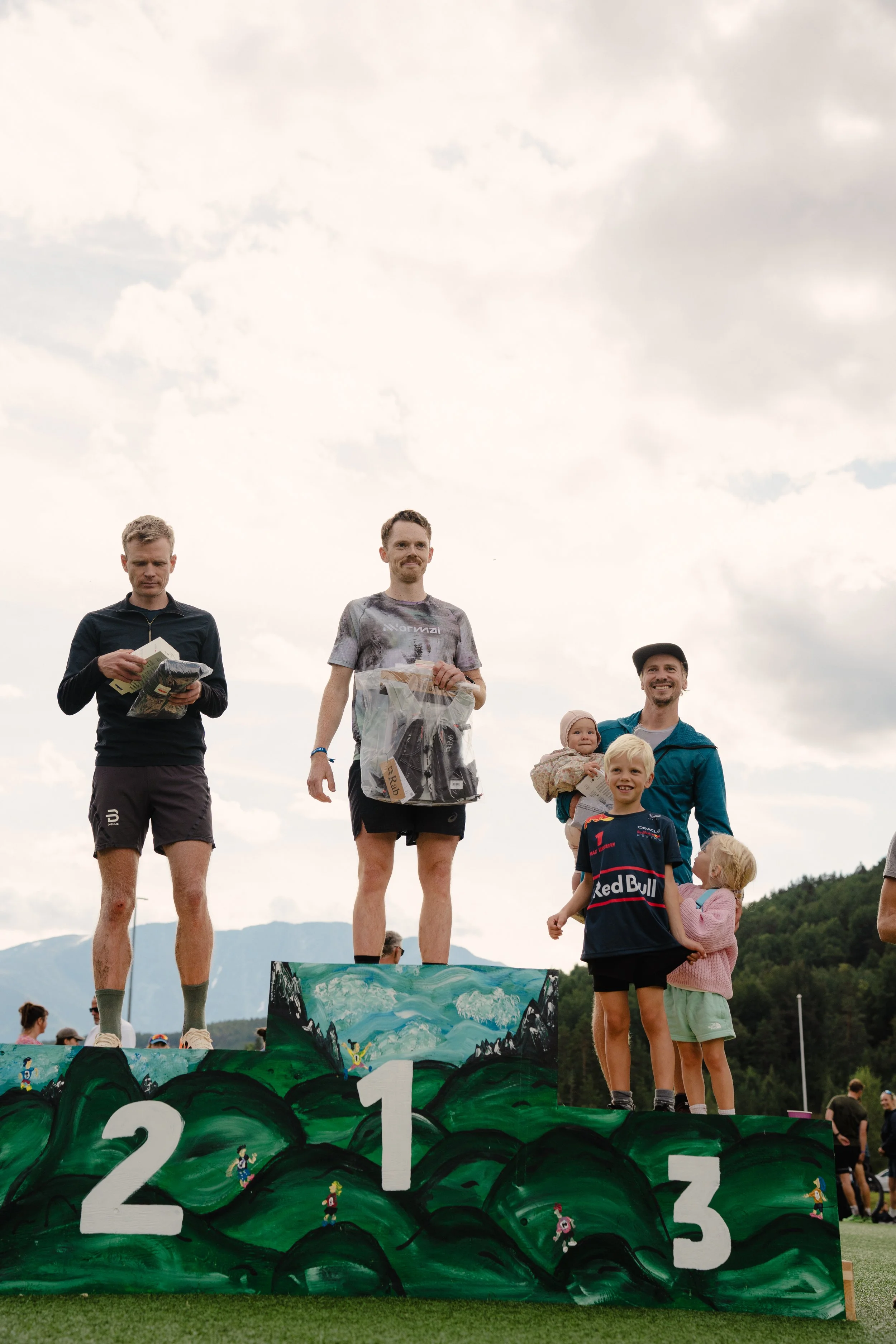 Three men standing on a winners' podium with mountains in the background, the middle man holding a plastic bag, and other people and children standing nearby. The podium is painted with a green landscape and numbered 1, 2, and 3.
