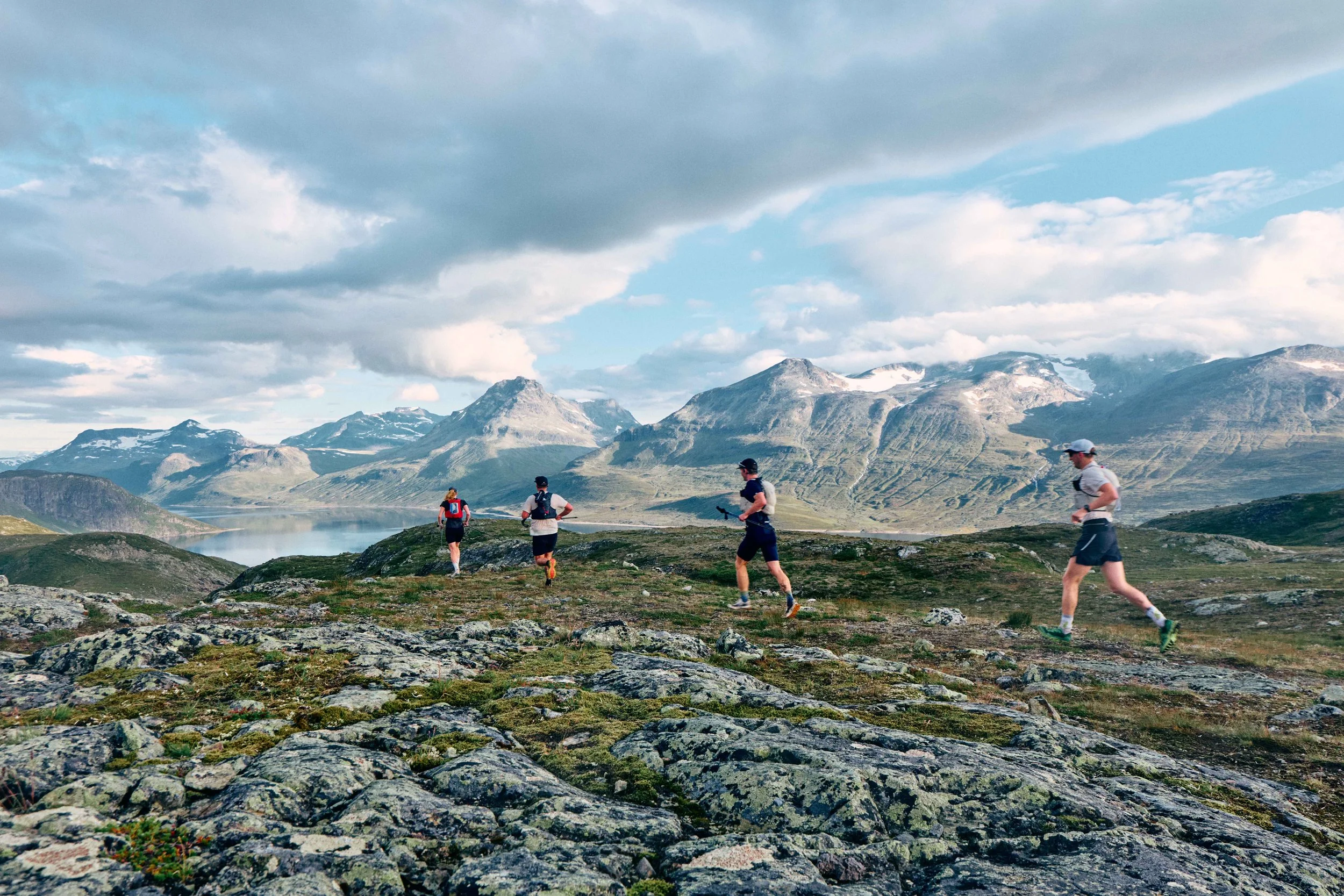 Four people trail running on rocky terrain in a mountainous landscape with snow-capped peaks, a lake, and partly cloudy sky in the background.