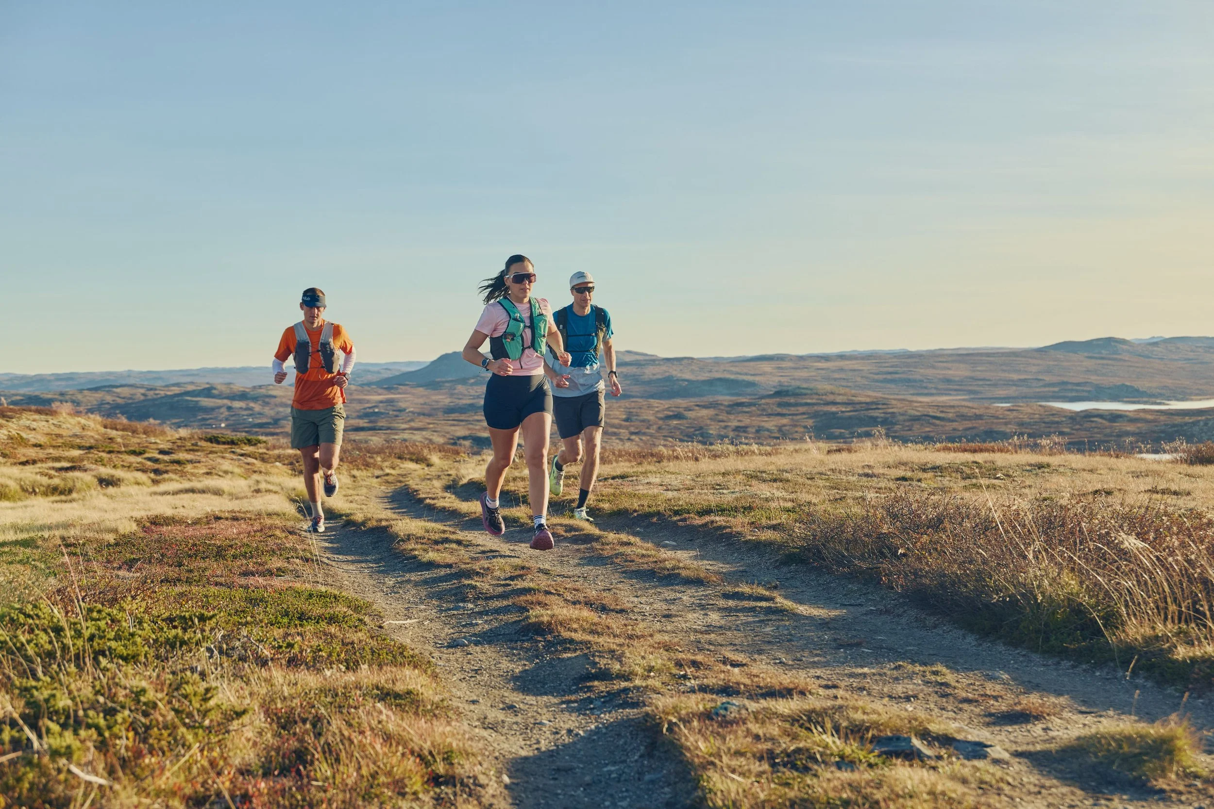 Three people trail running on a dirt path in an open grassy landscape with rolling hills in the background during daytime.