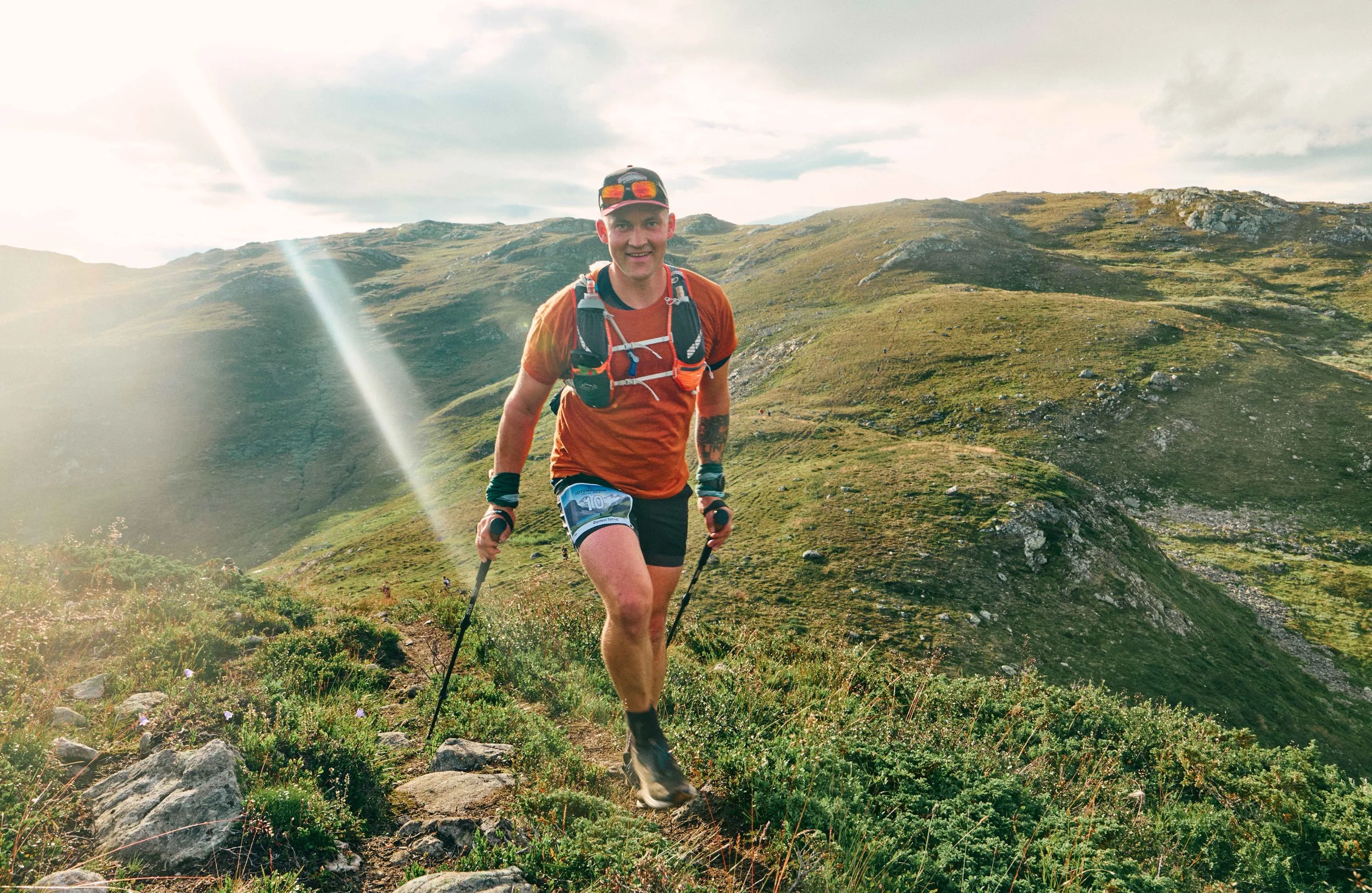 A man trail running on a mountain trail with rolling green hills in the background, wearing an orange shirt, black shorts, a hydration pack, and sunglasses.