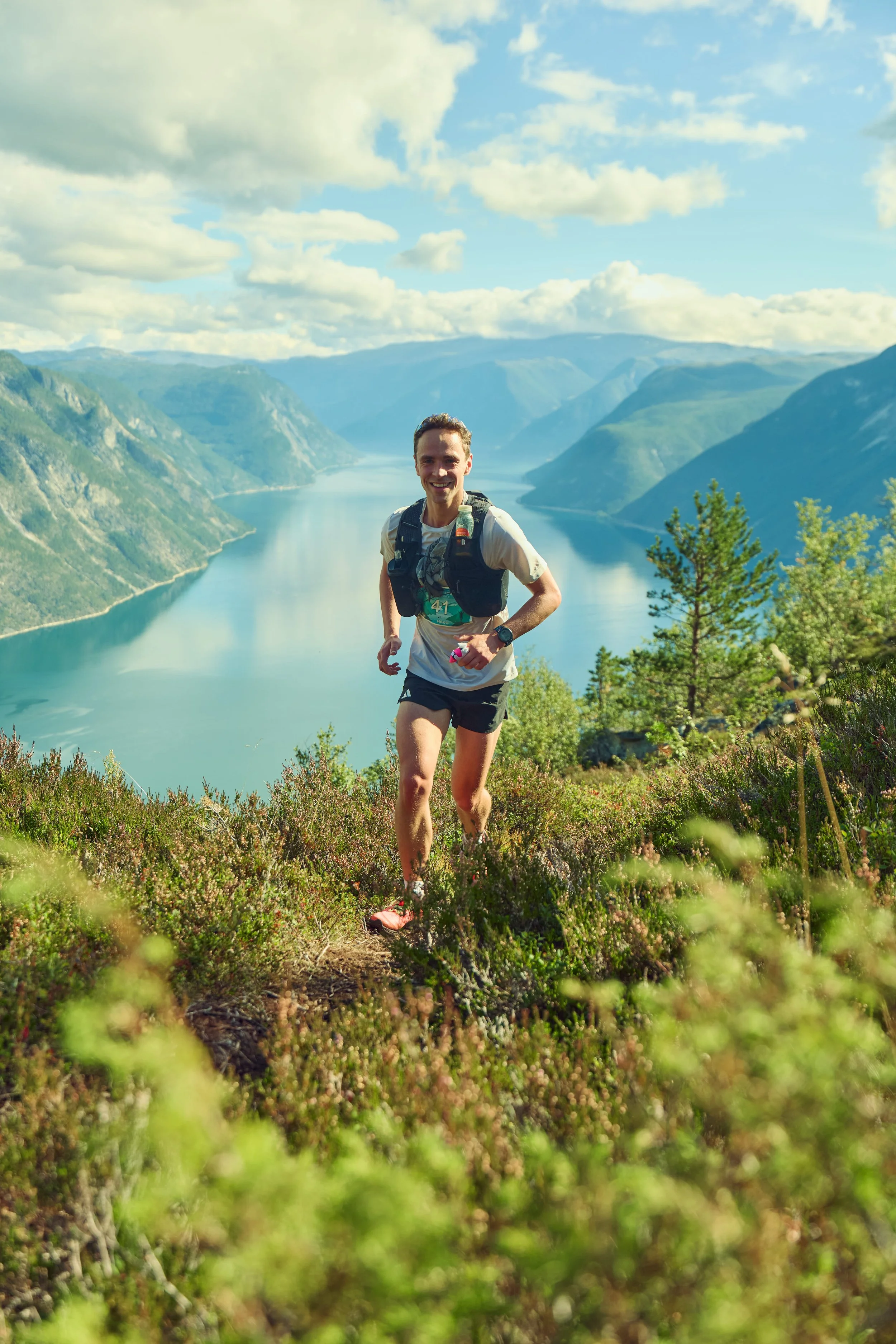 A man trail running on a mountain path overlooking a river in a canyon with mountains in the background, under a partly cloudy sky.
