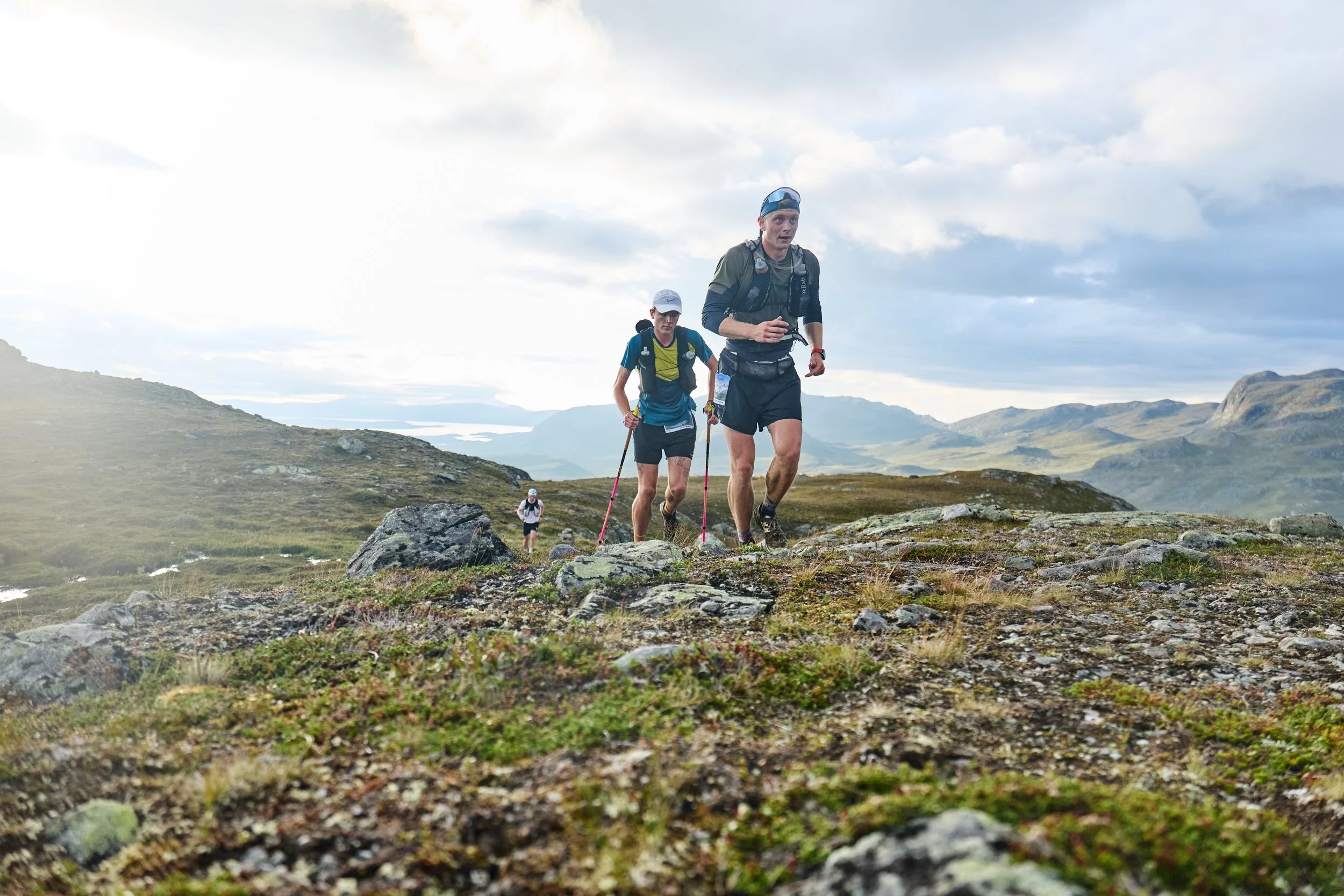 Three hikers trek across a rocky, grassy mountainous landscape under a partly cloudy sky.