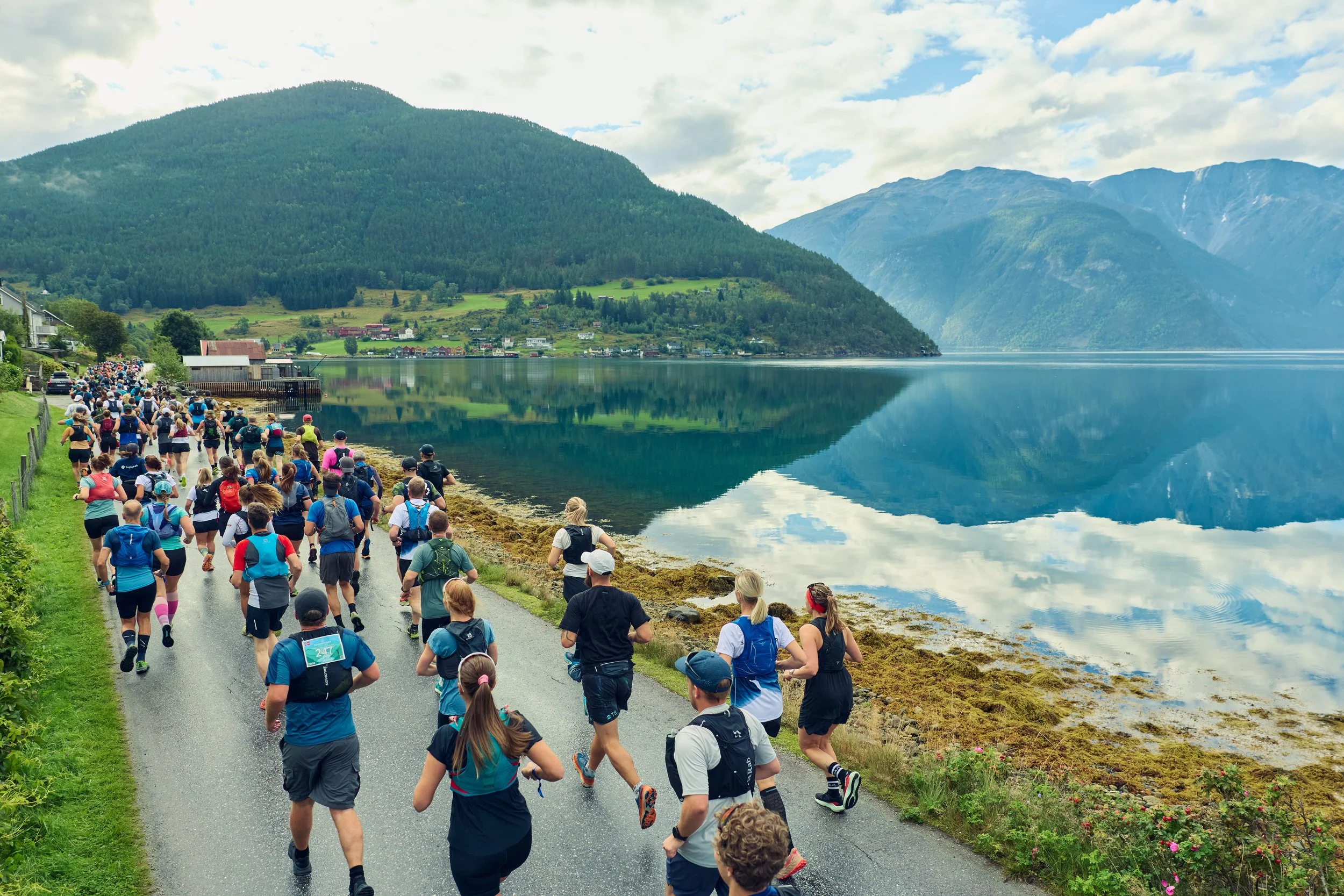 A large group of people participating in a marathon or race along a scenic lakeside path with mountains in the background.