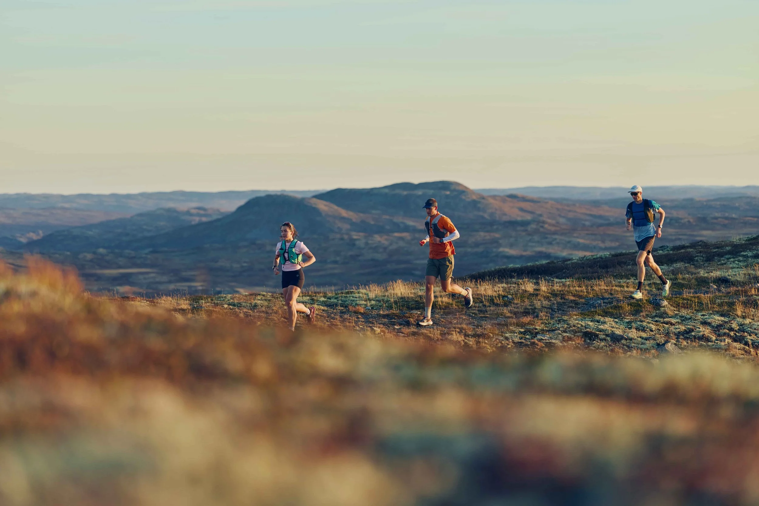 Three people trail running on a grassy hillside with mountains in the background during sunset.