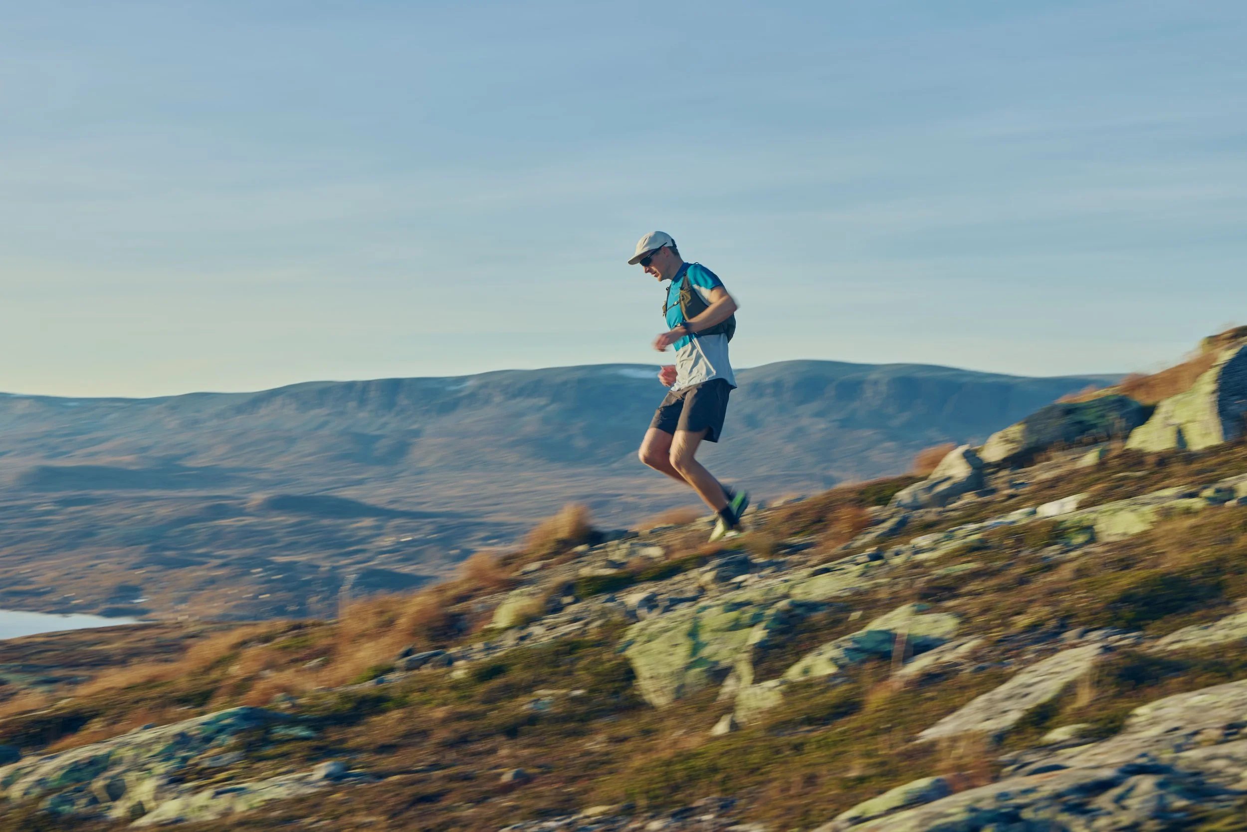 A man trail running on rocky terrain in a vast mountainous landscape during daytime.