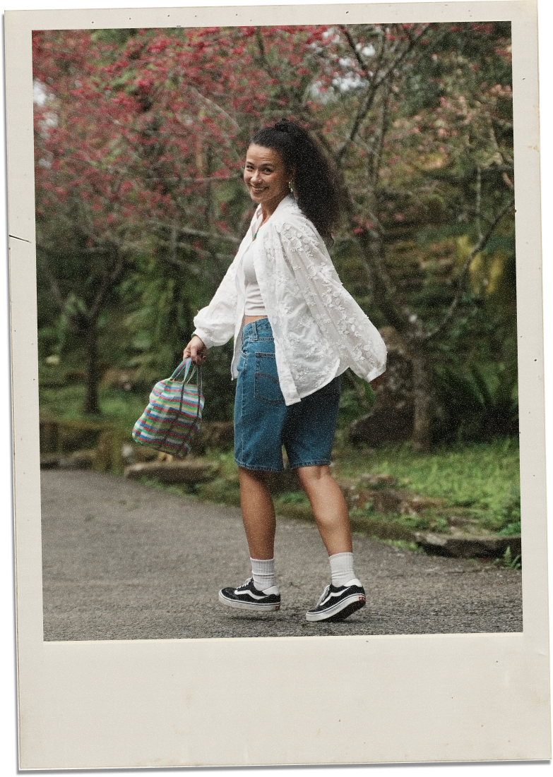 A woman standing outdoors on a paved path, smiling, with pink flowering trees and greenery in the background, carrying a colorful striped bag, wearing a white embroidered jacket, white crop top, denim shorts, white socks, and black Vans sneakers.