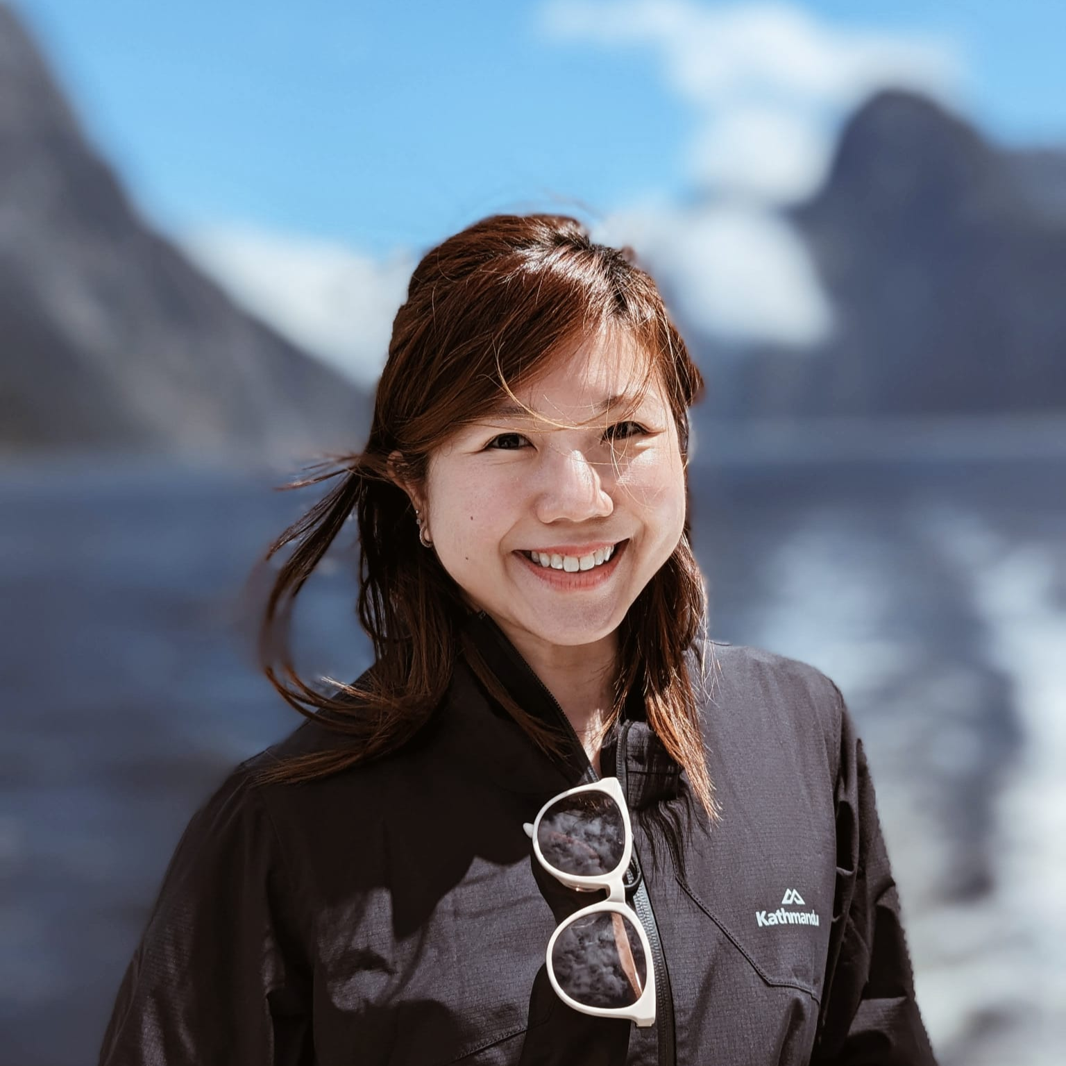 A woman smiling outdoors with mountains and blue sky in the background, wearing a black jacket with sunglasses hanging from the collar, and a Kathmandu logo on the jacket.