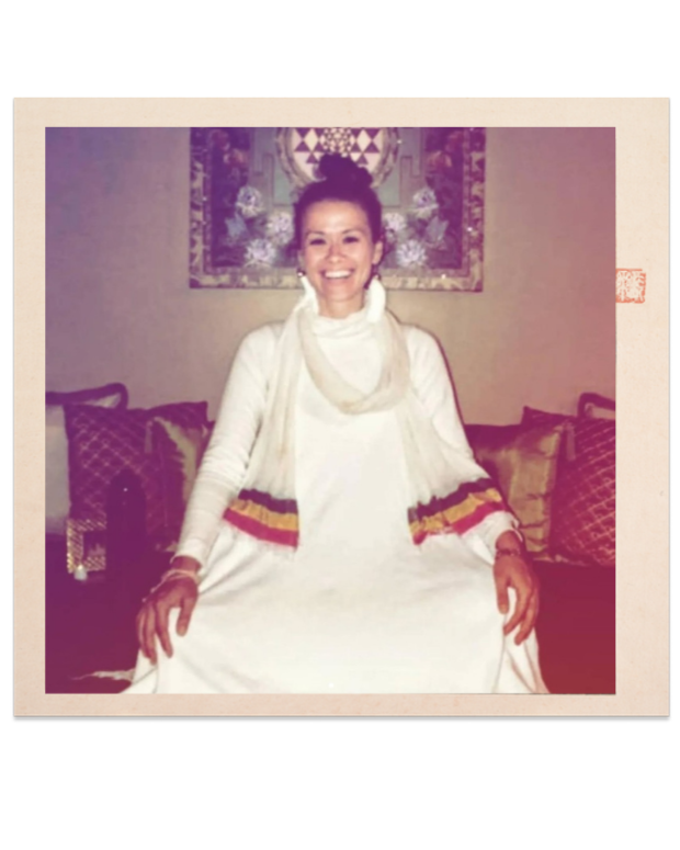 A woman sitting on a sofa, smiling, wearing a white dress with a cream-colored scarf, surrounded by decorative pillows, with artwork hanging on the wall behind her.