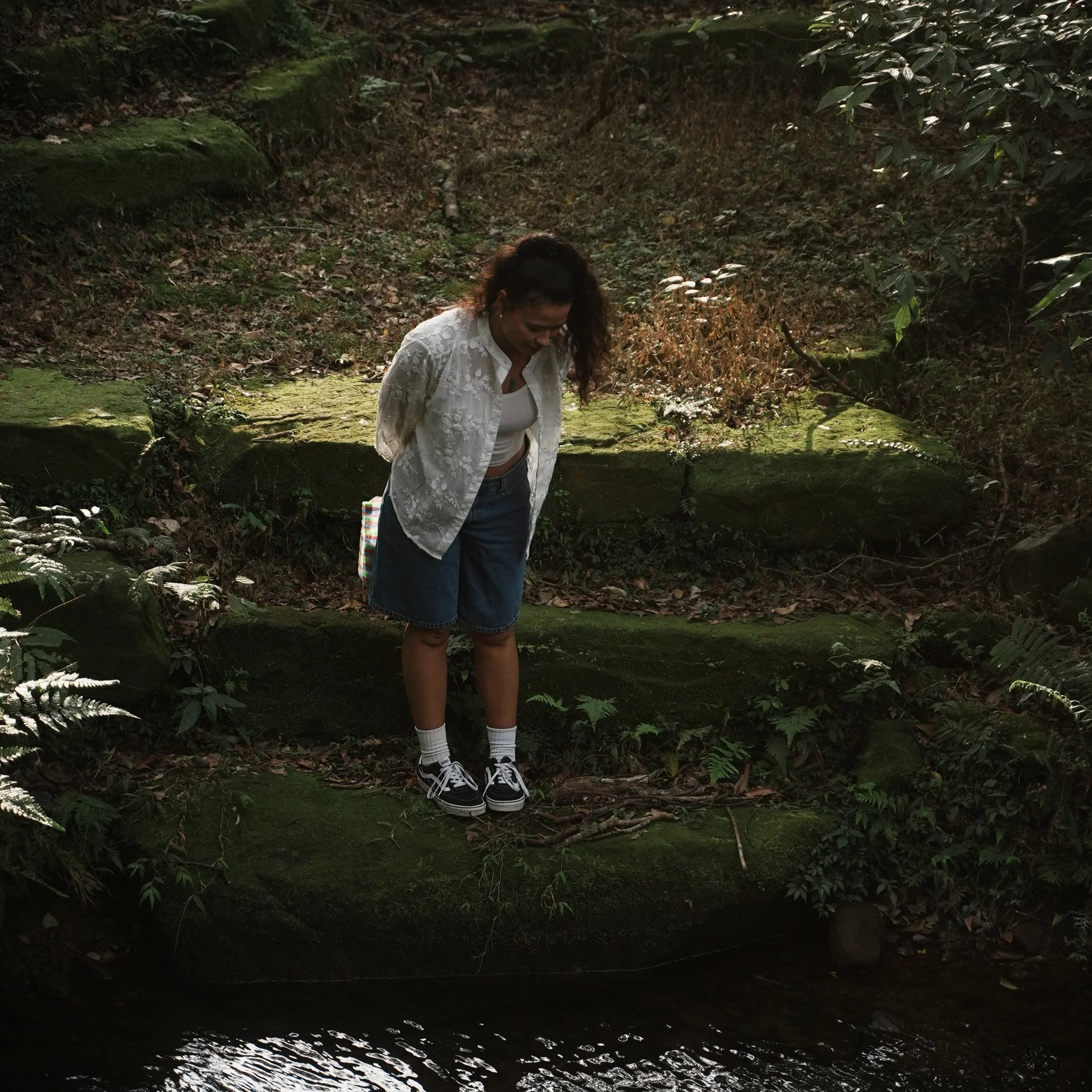A woman in a white jacket, denim skirt, and sneakers standing on grass in a park with a large tree in the background, smiling.