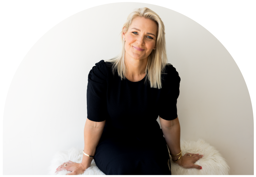 A woman with blonde hair, wearing a black dress, sitting on a white furry surface, smiling at the camera.