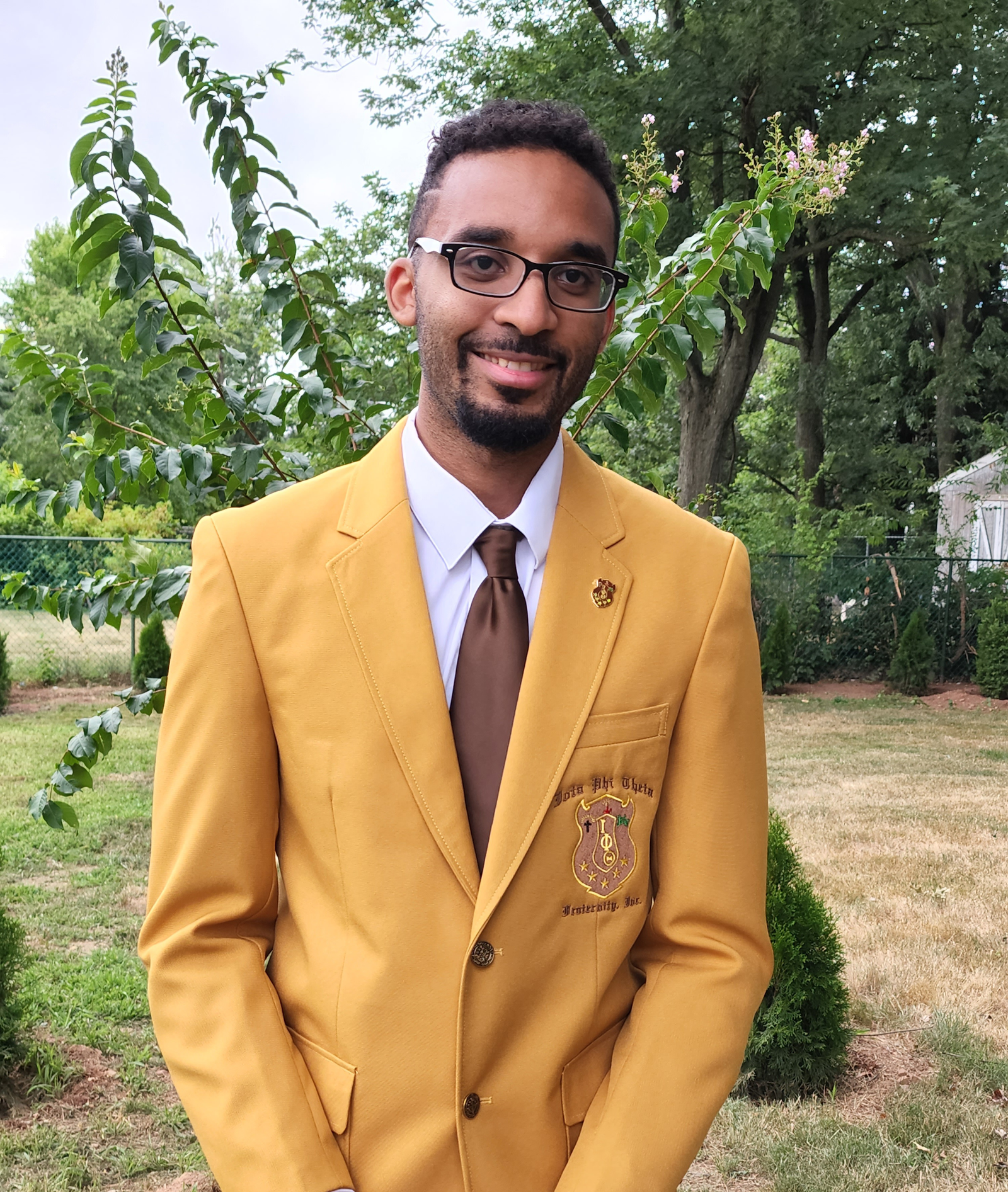 Portrait of a man wearing glasses and a yellow blazer with a crest on the pocket, standing outdoors with green trees in the background.