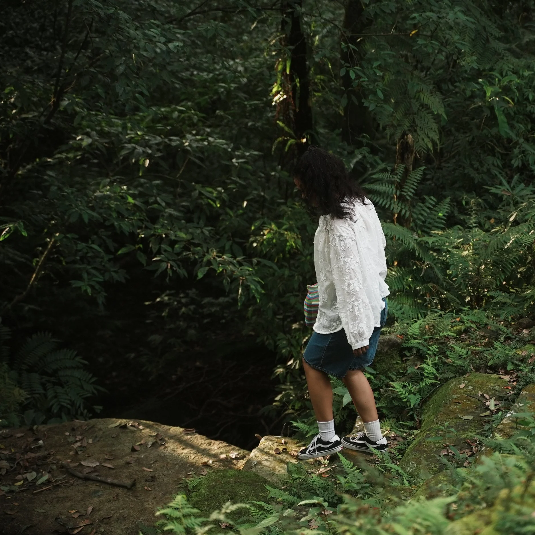 Young girl walking outdoors on green grass, carrying a striped bag, wearing a white jacket, denim shorts, and sneakers, with trees in the background.