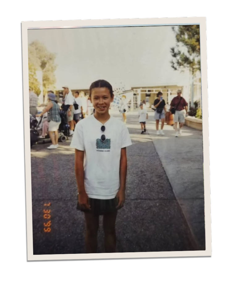 A young girl with dark hair tied back, smiling and standing outside on a sunny day in a crowded area, wearing a white T-shirt, a dark skirt, and sunglasses hanging from her shirt.