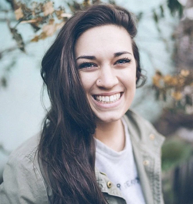 Young woman with long dark hair smiling outdoors in front of a blurred background of leaves.