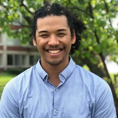 A smiling man with curly dark hair and a beard, wearing a light blue button-up shirt, standing outdoors near a tree with green leaves.