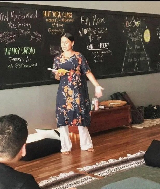 A woman standing on a wooden floor in front of a blackboard with colorful writing, holding a book, with an audience in the foreground. The blackboard lists various classes and events including Hula Yoga Class, Hip Hop Cardio, Full Moon Women's Circle, and Kids Camp.