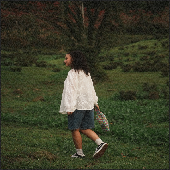 Young girl walking outdoors on green grass, carrying a striped bag, wearing a white jacket, denim shorts, and sneakers, with trees in the background.
