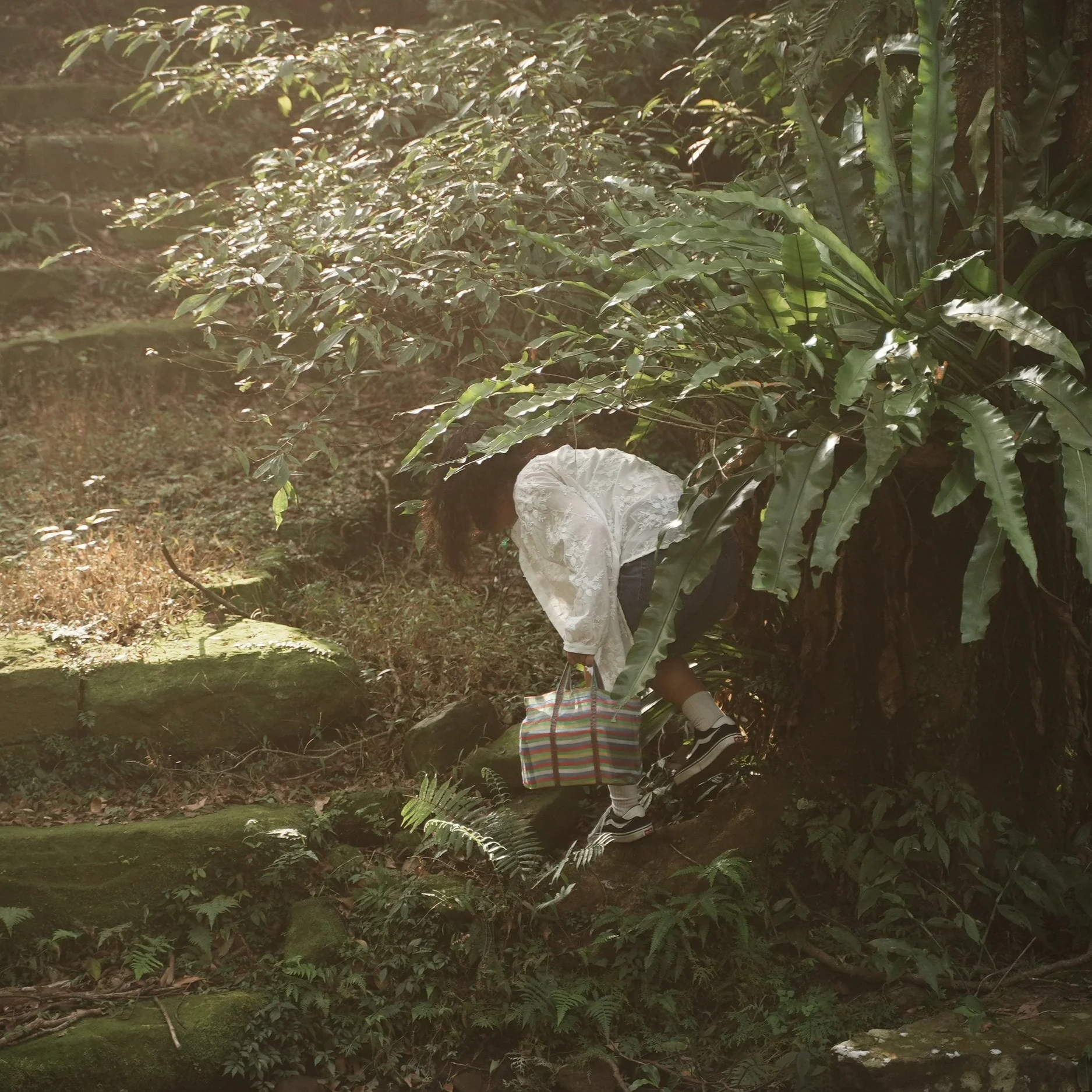 A woman with curly hair, wearing a white blouse, denim skirt, and sneakers, carrying a colorful striped bag, standing on grass in a park or garden with trees in the background.