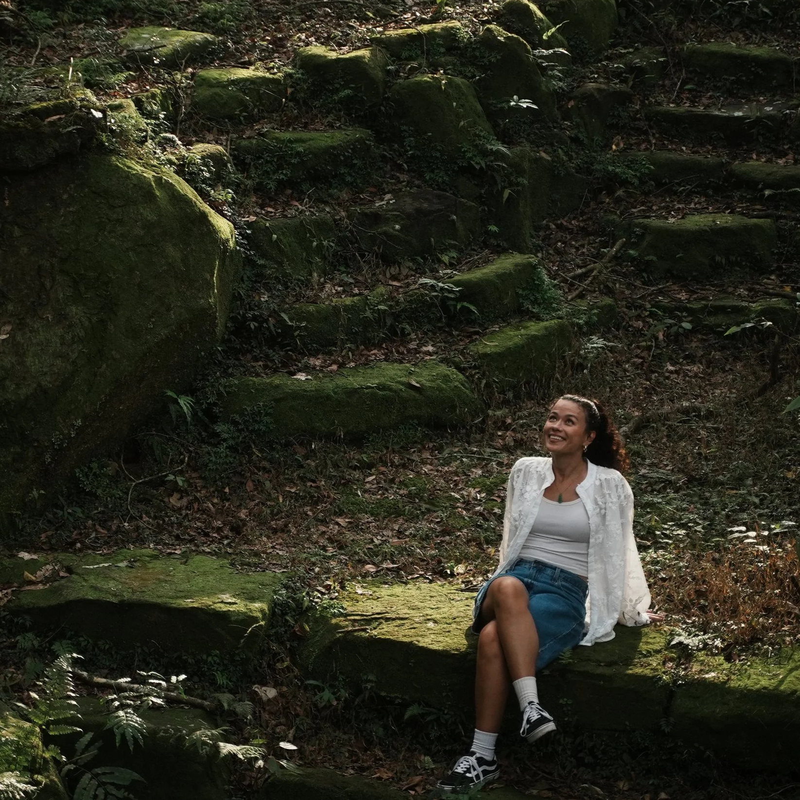 A girl wearing a white sweater, blue skirt, and white shoes walking on a grassy field with trees in the background.