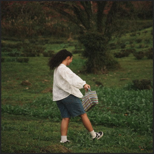 A woman with curly hair, wearing a white blouse, denim skirt, and sneakers, carrying a colorful striped bag, standing on grass in a park or garden with trees in the background.