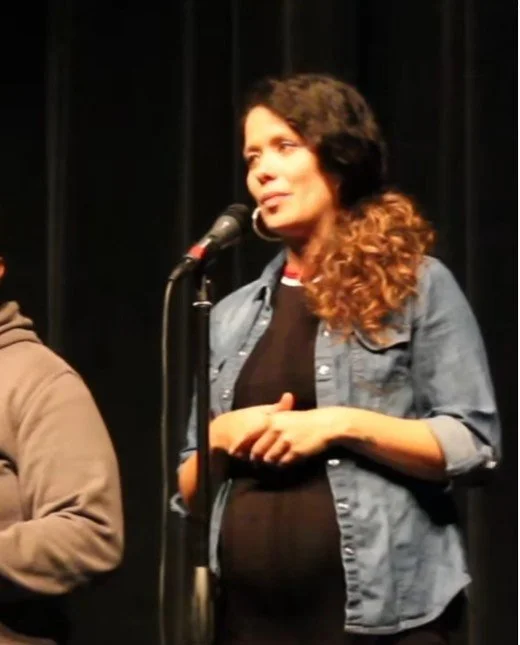 A woman with wavy hair speaking into a microphone on stage, wearing a denim jacket over a black shirt, with a black curtain in the background.