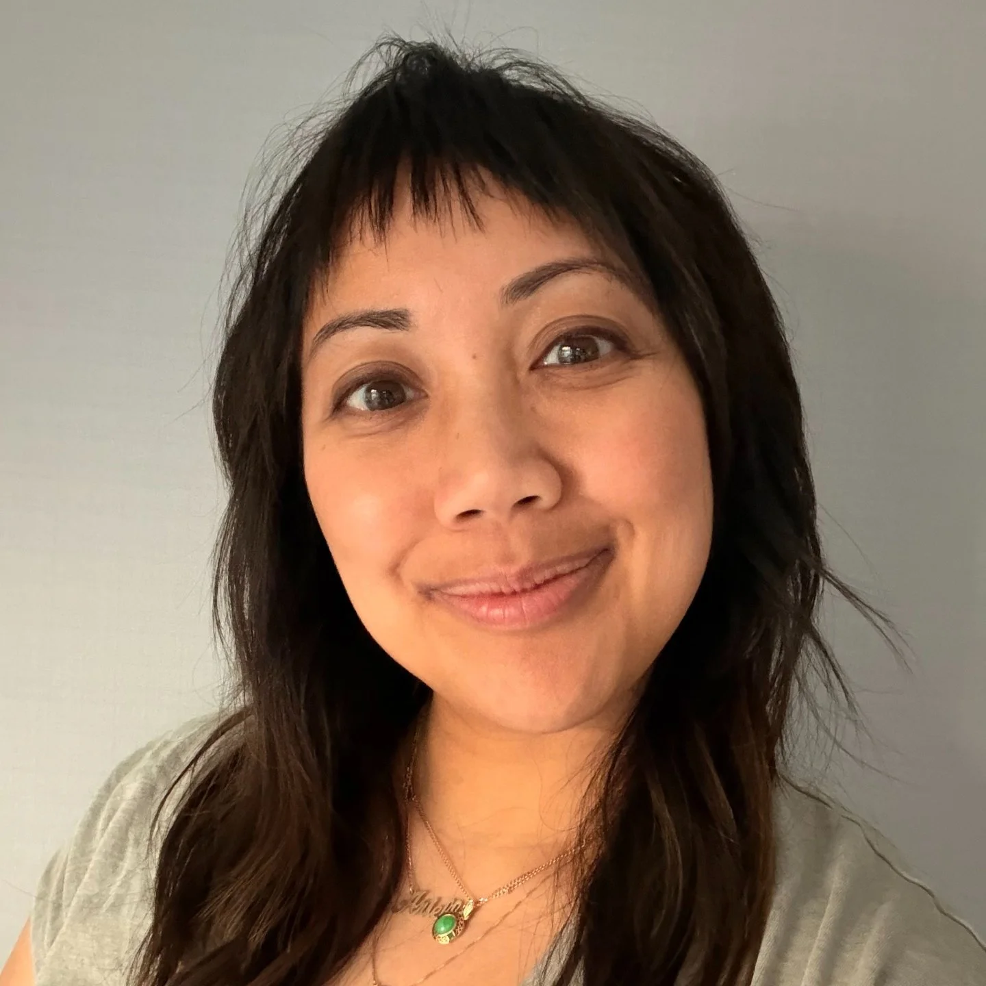 Close-up of a smiling woman with medium-length dark hair, wearing a light-colored shirt and a gold necklace with a green pendant, standing against a plain neutral background.