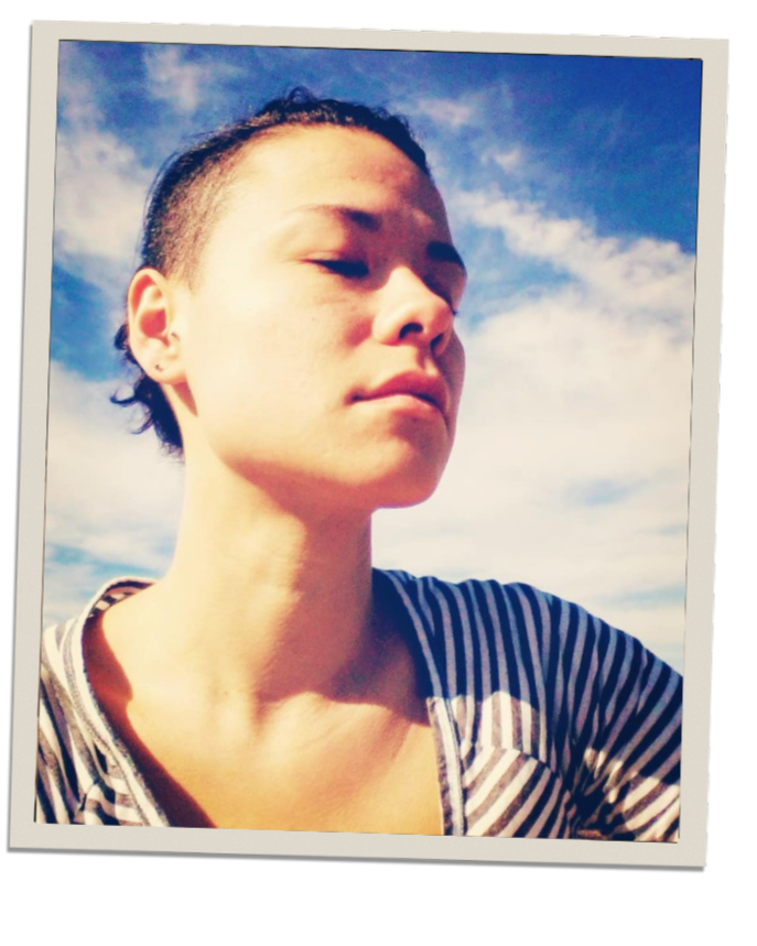A person with short, dark, slicked-back hair taking a selfie outdoors against a bright blue sky with some clouds, wearing a striped shirt.
