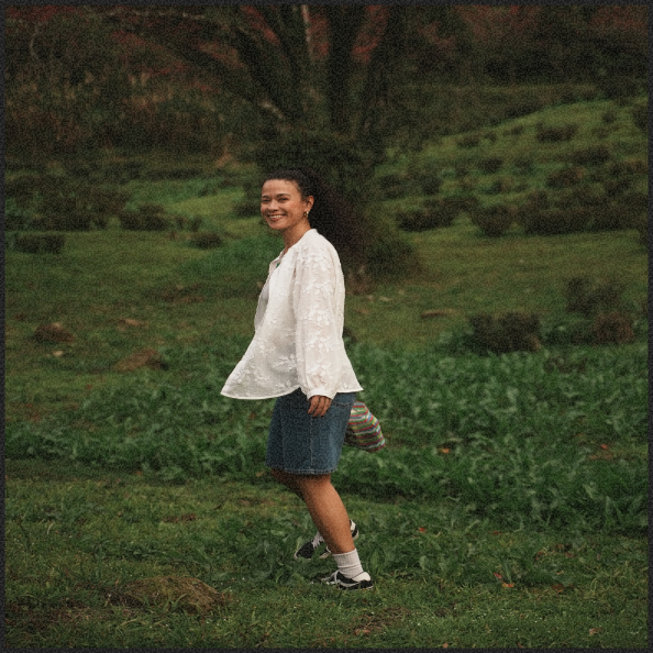 A woman in a white jacket, denim skirt, and sneakers standing on grass in a park with a large tree in the background, smiling.