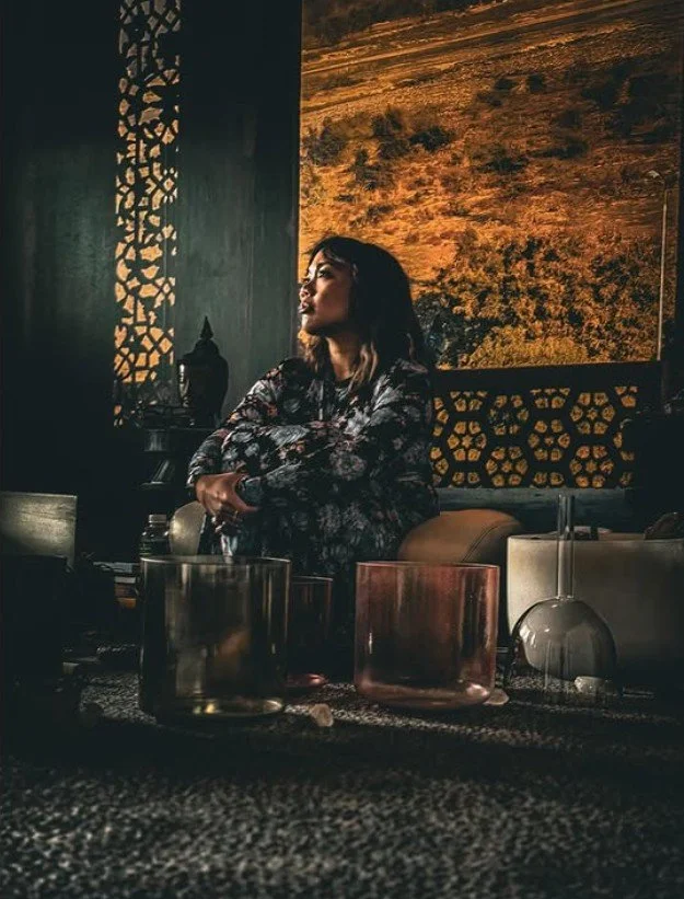 A beige, speckled ceramic vase with a dried plant inside, placed on a wooden surface with sunlight casting shadows on a plain wall.
