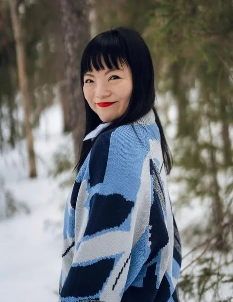 A woman with black hair and red lipstick smiling outdoors in a snowy forest, wearing a blue and white patterned sweater.
