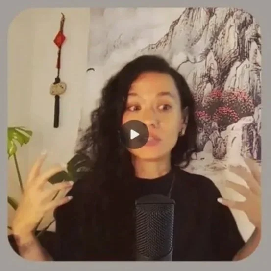 A woman with curly dark hair gesturing with her hands while speaking into a microphone in a room decorated with a mountain landscape, pink flowers, a hanging ornament, and a plant.