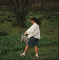 A girl wearing a white sweater, blue skirt, and white shoes walking on a grassy field with trees in the background.