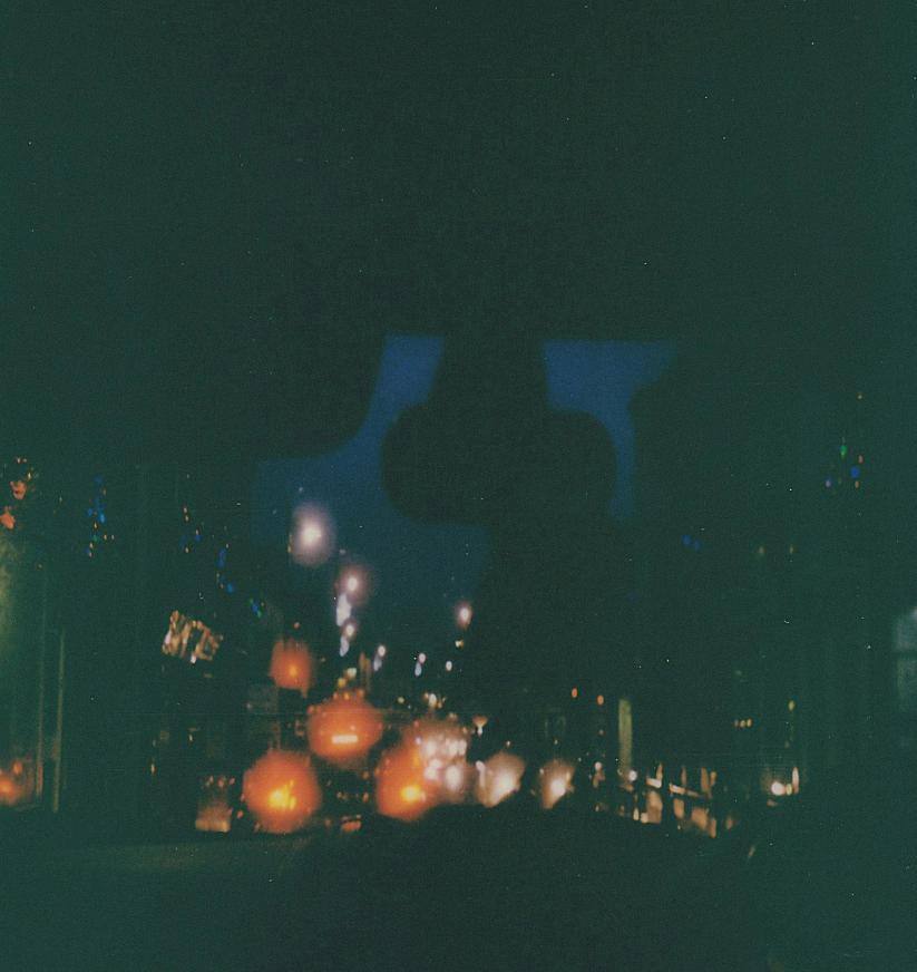 Nighttime city street with blurred lights and reflections, viewed from inside a vehicle.