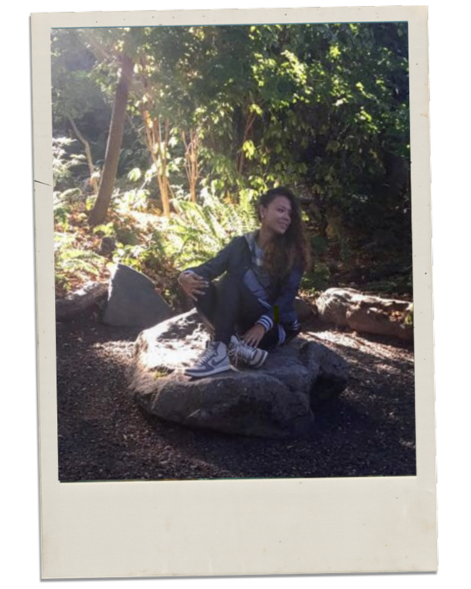 A girl sitting on a large rock in a wooded area, surrounded by trees and ferns, with sunlight filtering through the leaves.