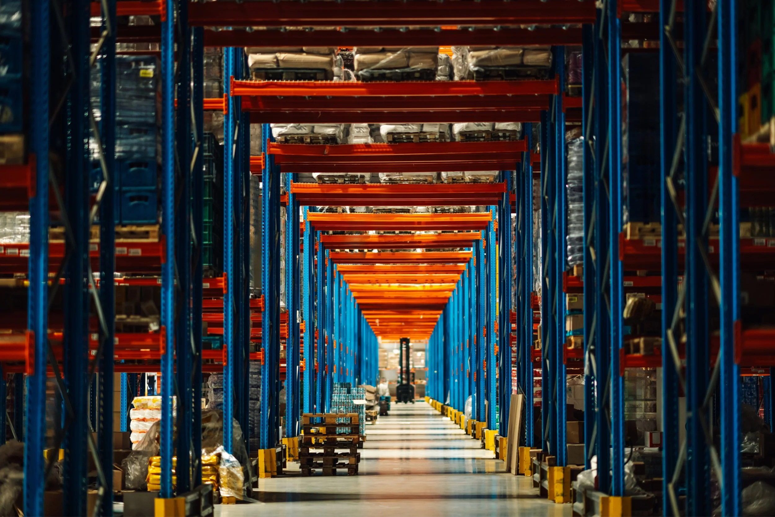 Wide view of tall warehouse shelves with orange beams and blue support frames, stocked with various goods, and a forklift in the distance.
