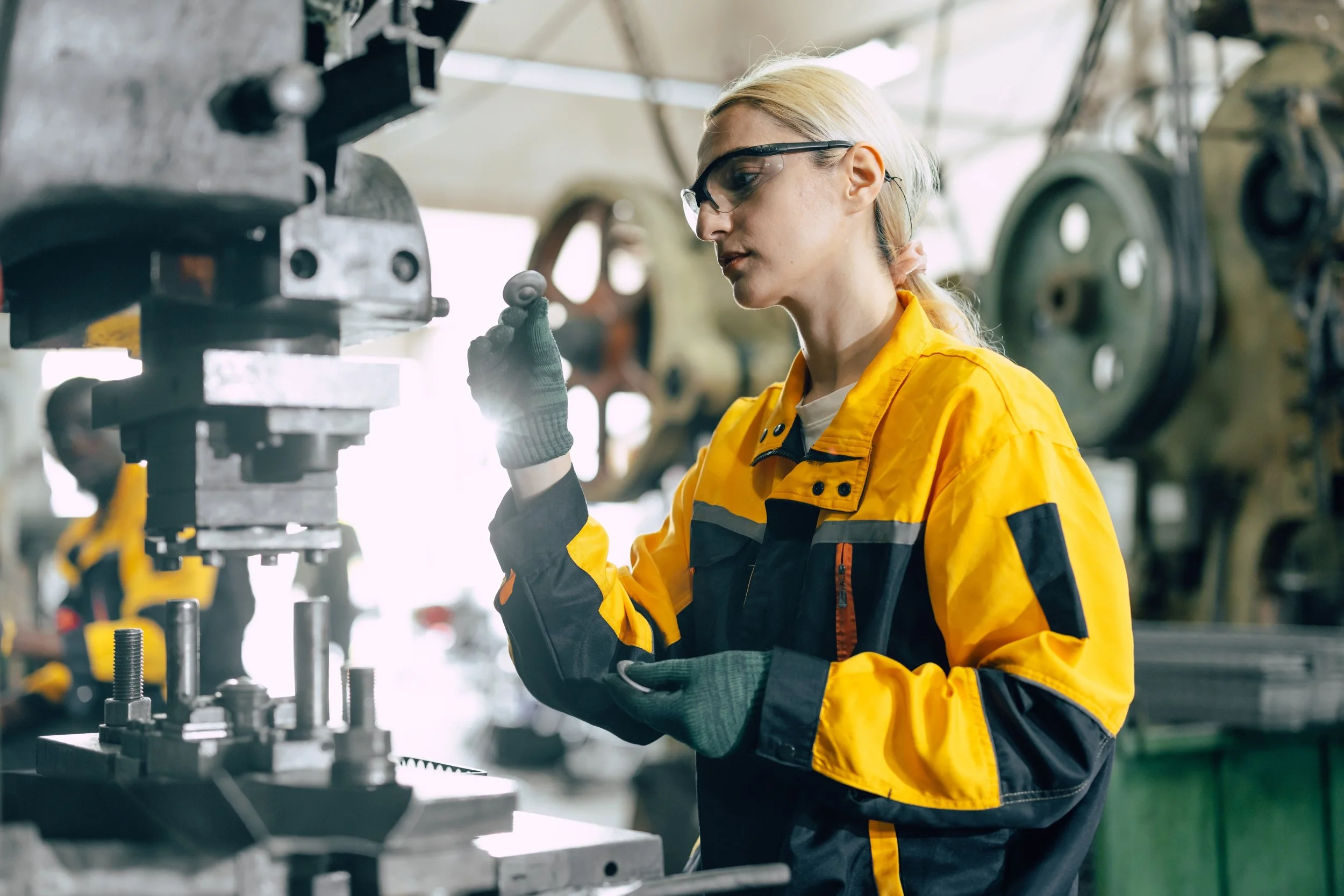 A woman in safety glasses, gloves, and a yellow and black work uniform inspecting machinery in a factory or workshop.