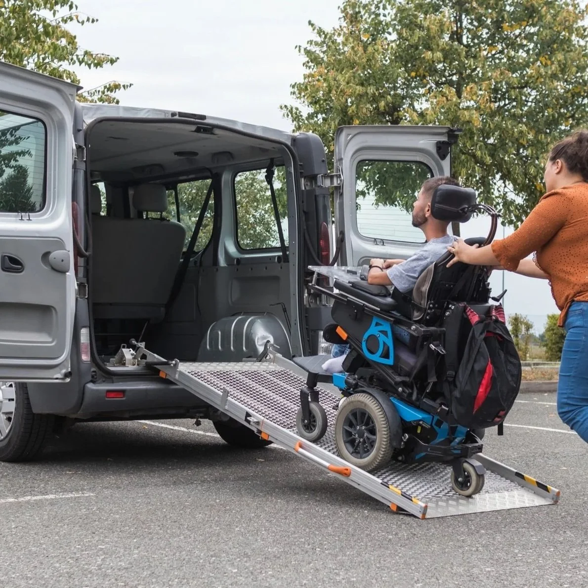 A person in a motorized wheelchair being guided into a van via a wheelchair lift, with a woman assisting.