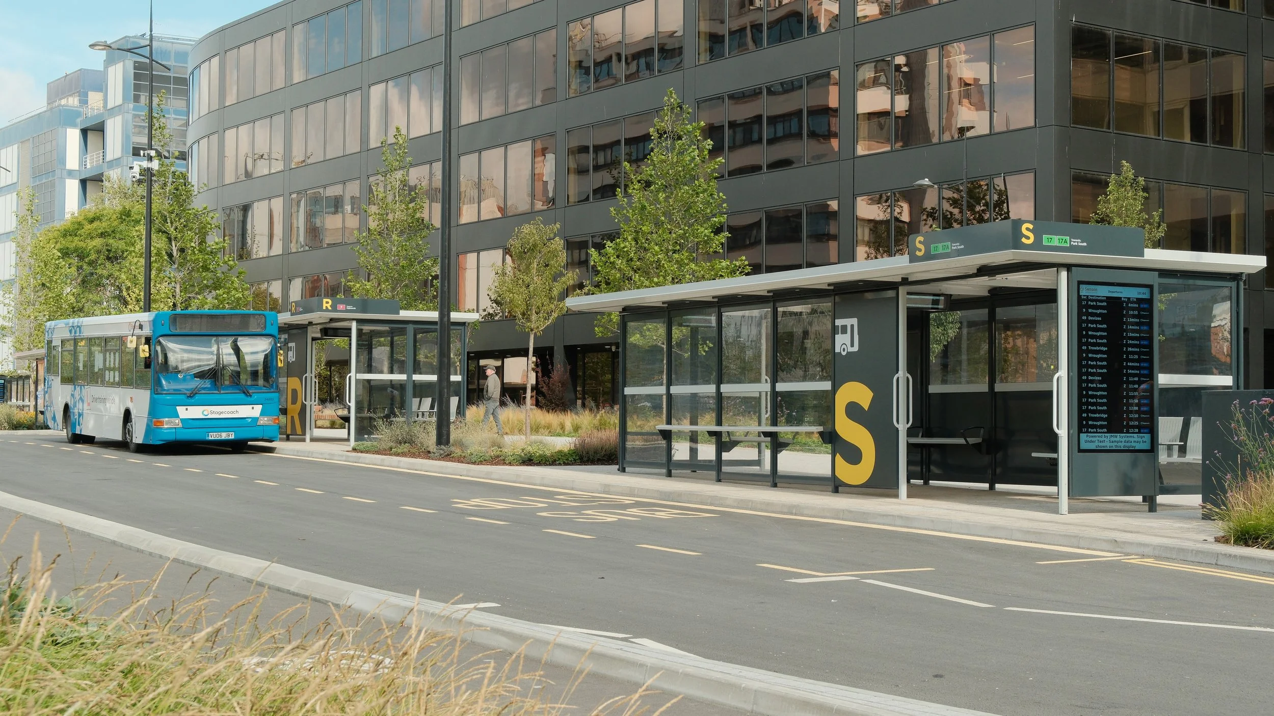 Modern city bus stop with shelters, digital timetable displays, a blue electric bus, and a high-rise office building in the background.