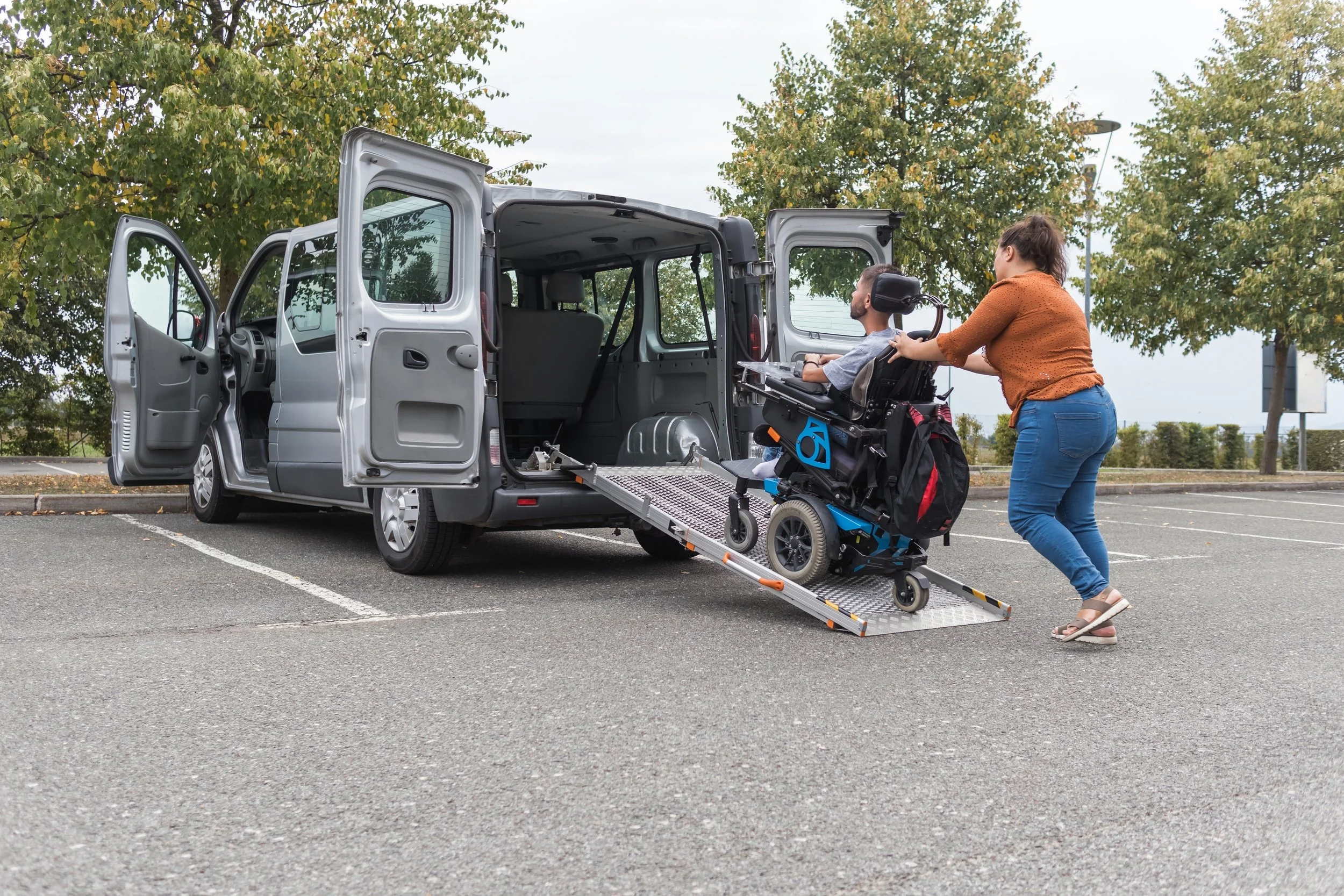 A woman helps a man in a motorized wheelchair onto a vehicle through an accessible ramp in a parking lot with trees in the background.
