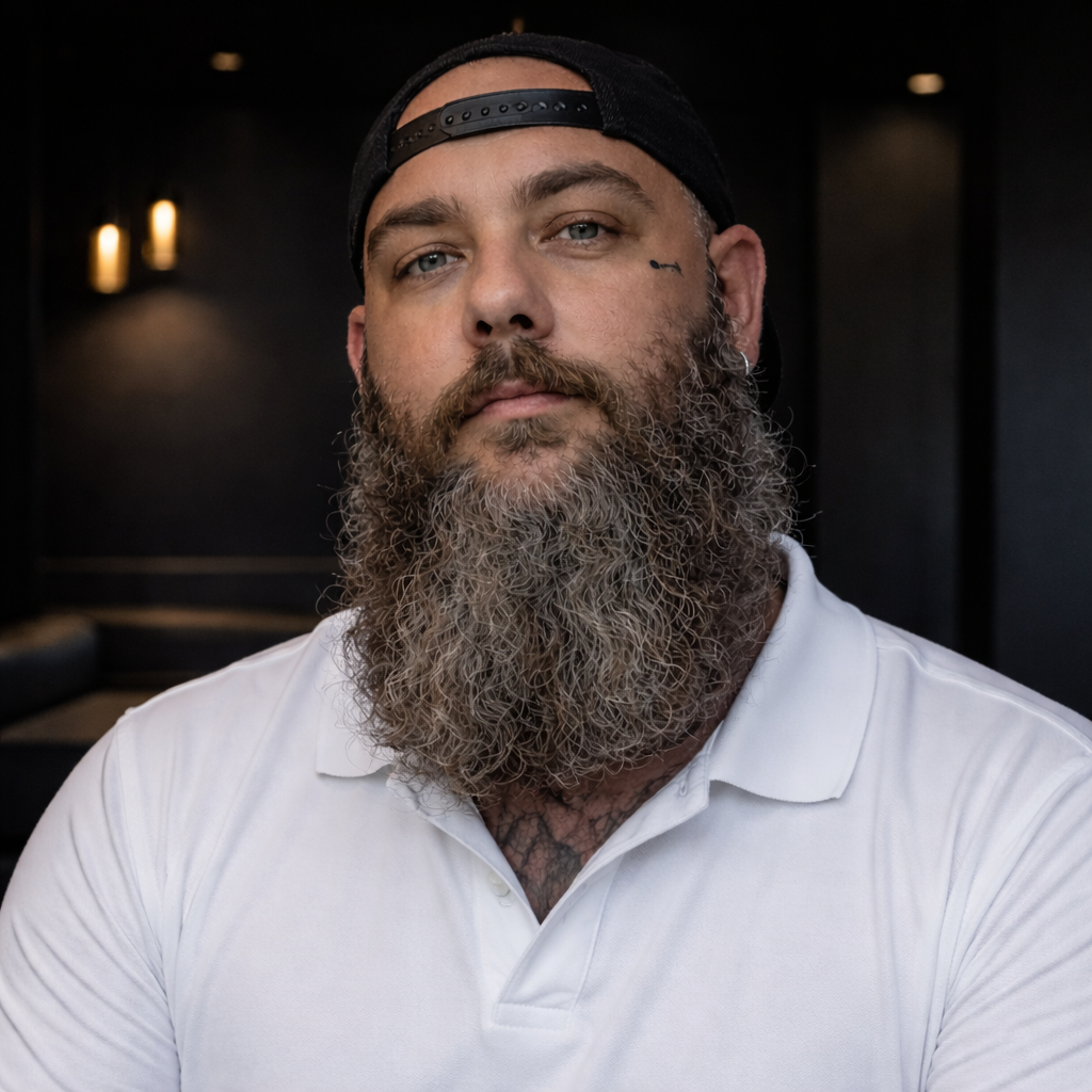A man with a long beard wearing a white polo shirt and a backwards black cap, looking into the camera with a serious expression, in a dark indoor setting.