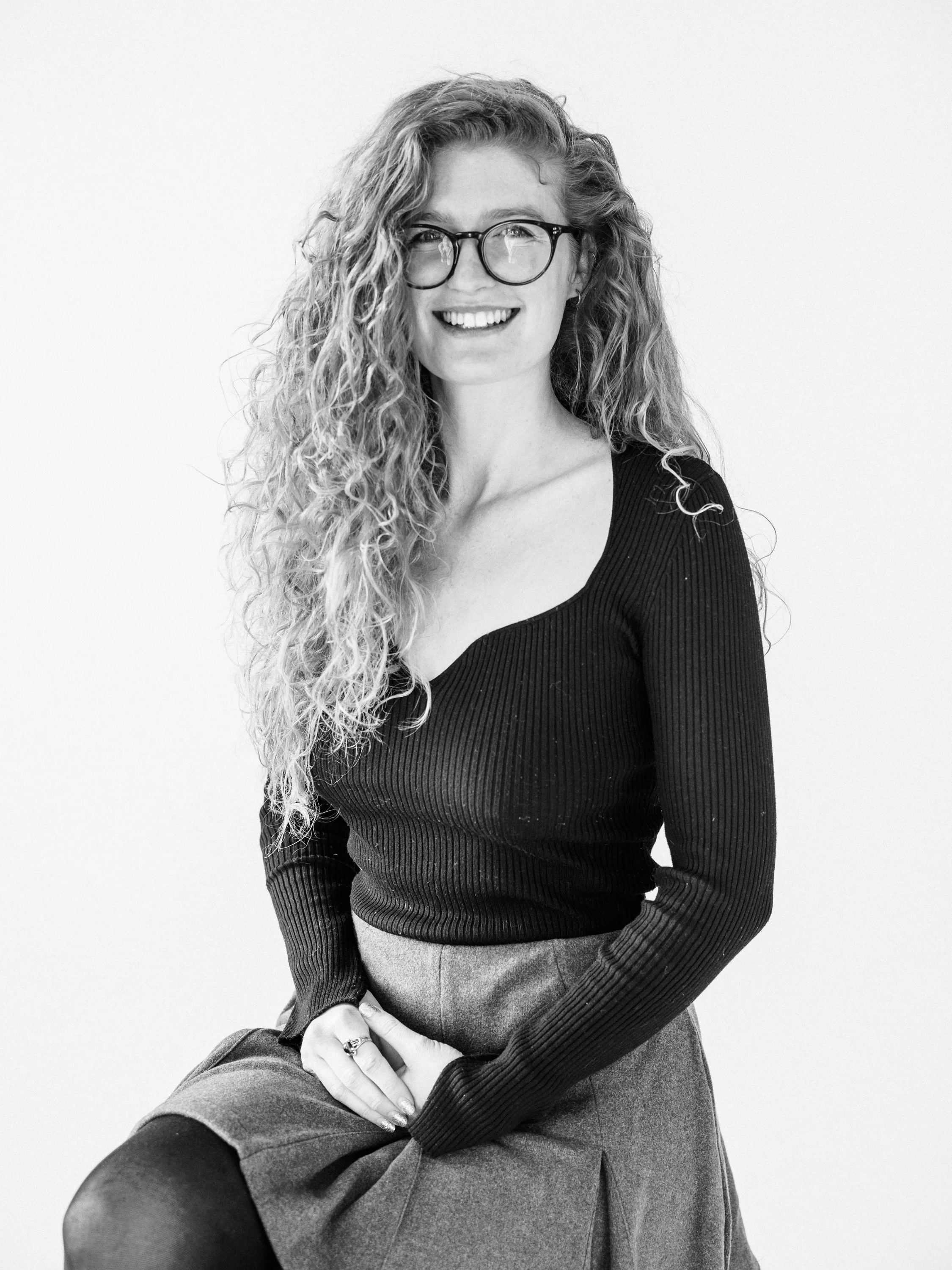Black and white portrait of a woman with curly hair, wearing glasses, a long-sleeved top, and a skirt, smiling and sitting against a plain background.