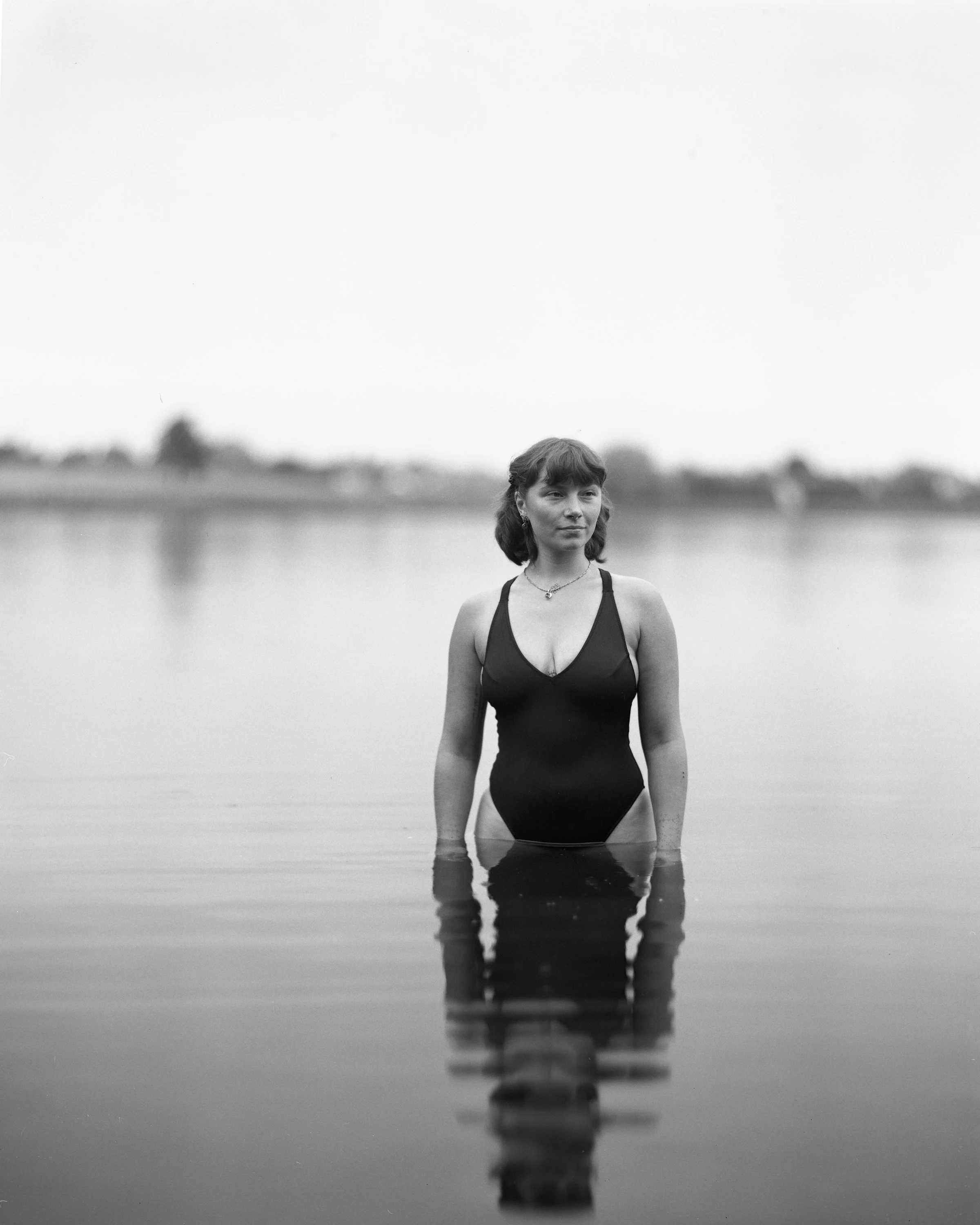 Woman standing in water at a lake, wearing a black swimsuit, with a calm expression, black and white photograph.