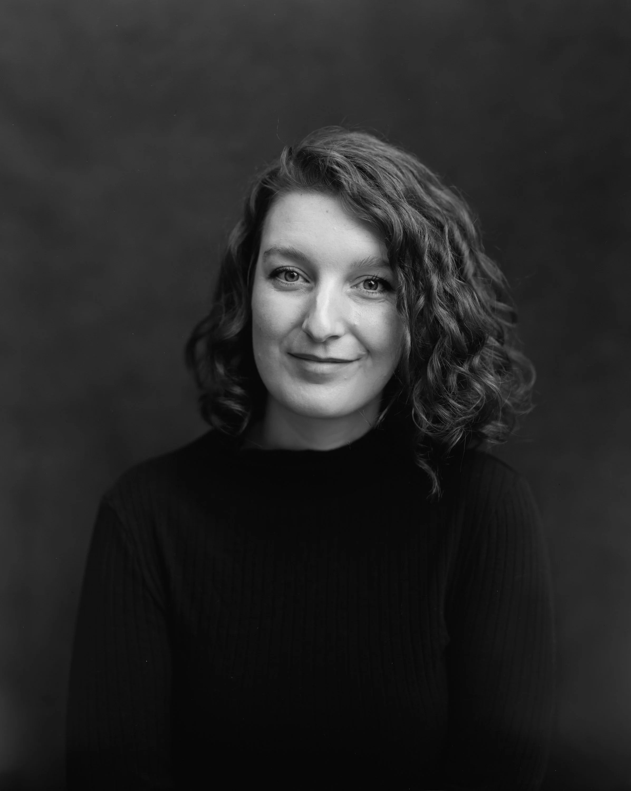 Black and white portrait of a woman with shoulder-length curly hair wearing a dark top, smiling gently, against a plain background.