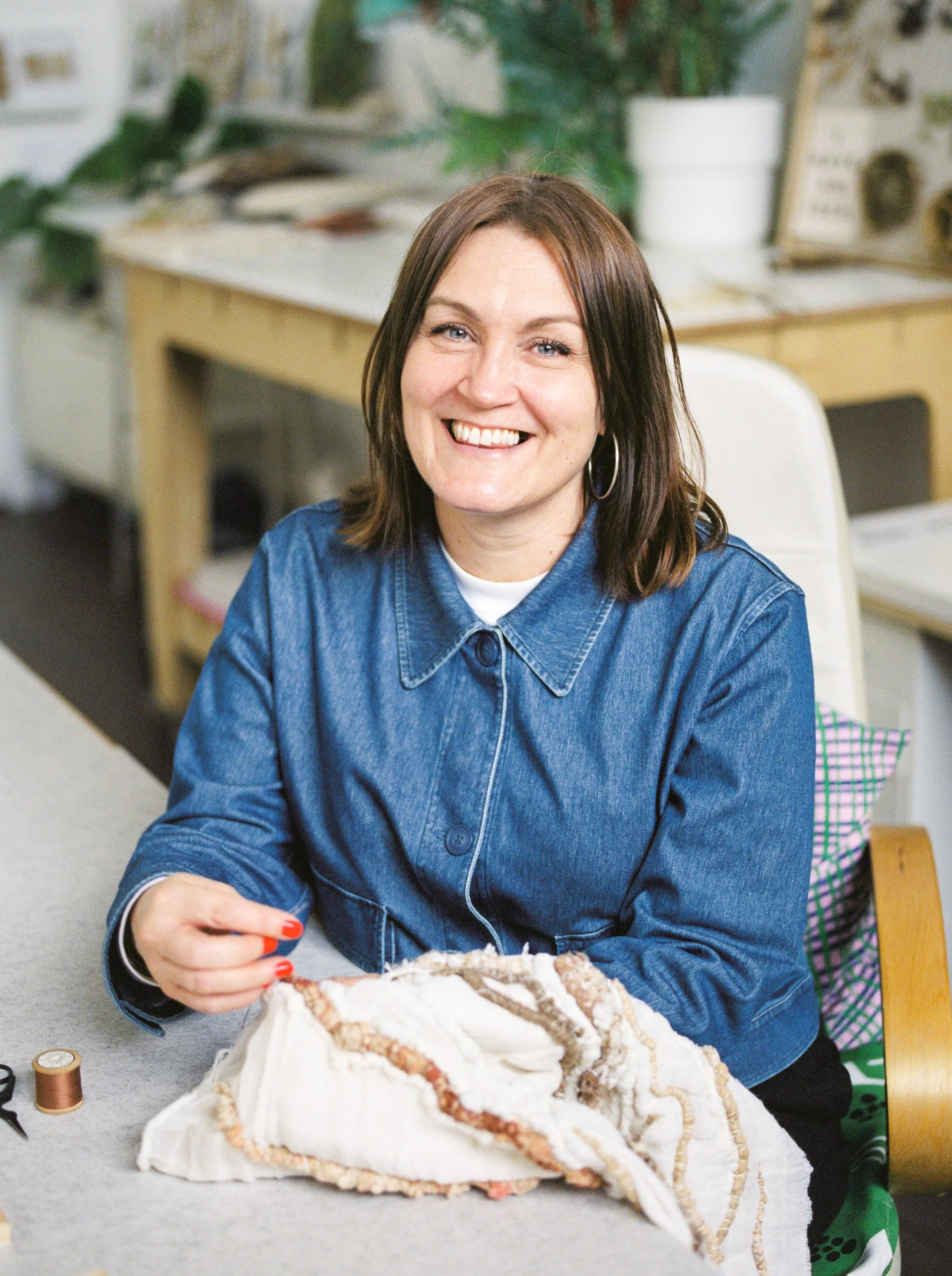 A woman with brown hair and earrings sitting at a table, smiling, working on a fabric project, with sewing supplies nearby in a cozy workspace.