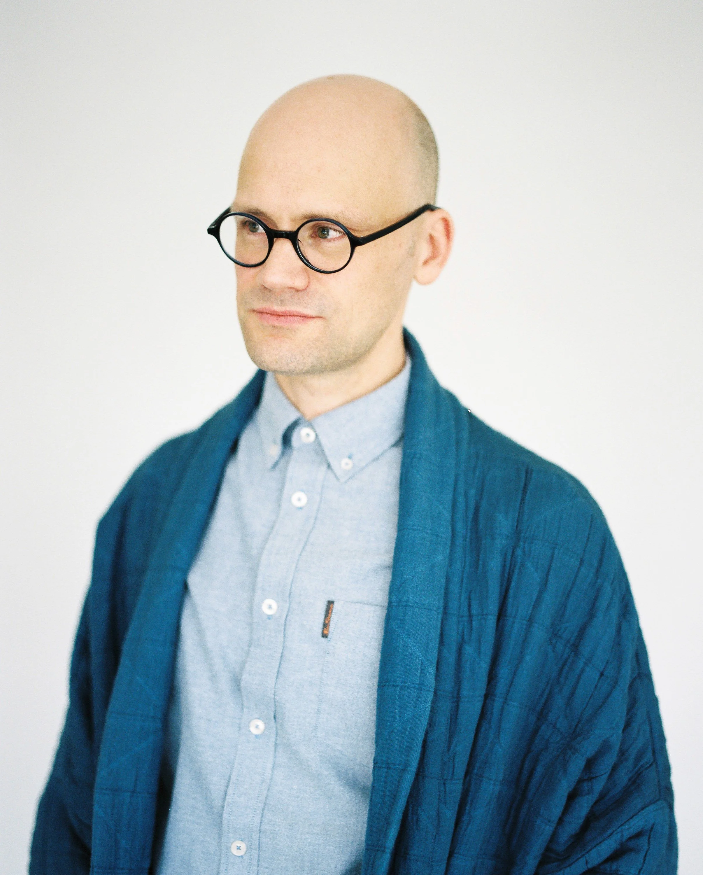 Portrait of a bald man wearing glasses, a light blue shirt, and a dark blue jacket, standing against a plain white background.