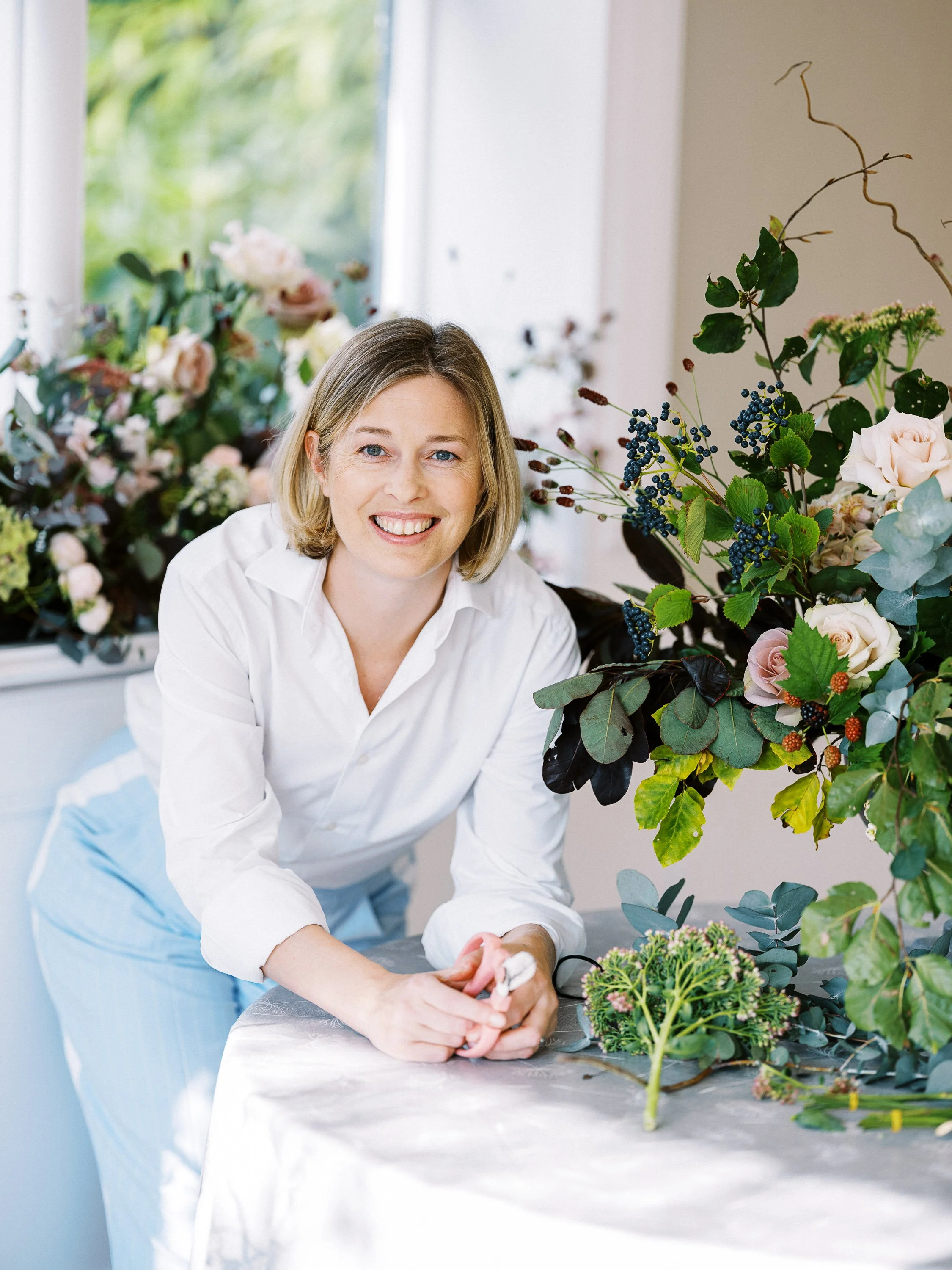 A woman smiling and working on floral arrangements in a bright space with large windows, surrounded by various flowers and greenery.