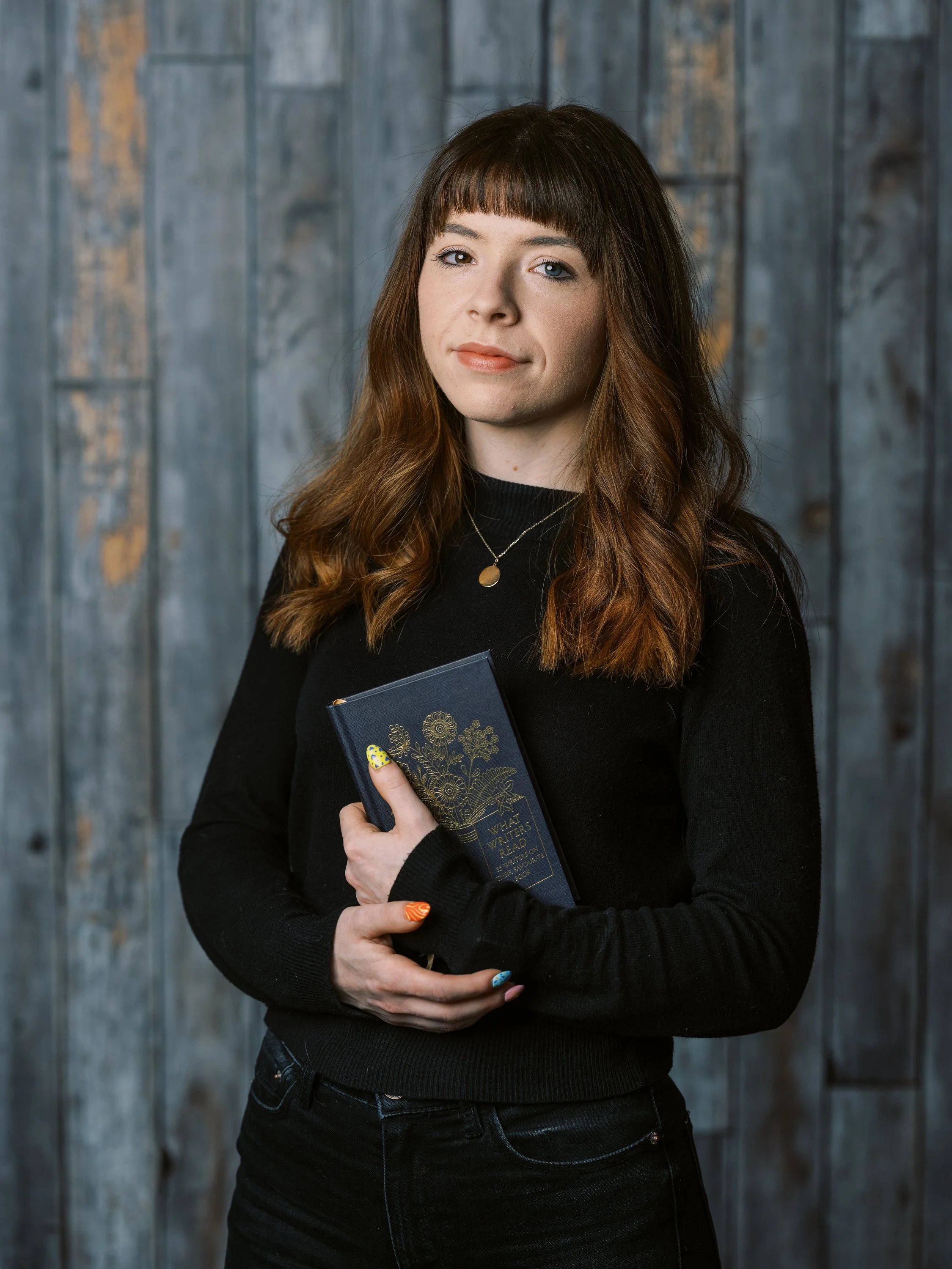 A young woman with long, wavy brown hair and bangs, wearing a black sweater and holding a book titled "What Writers Read." She is standing in front of a wooden wall with blue and gray tones.