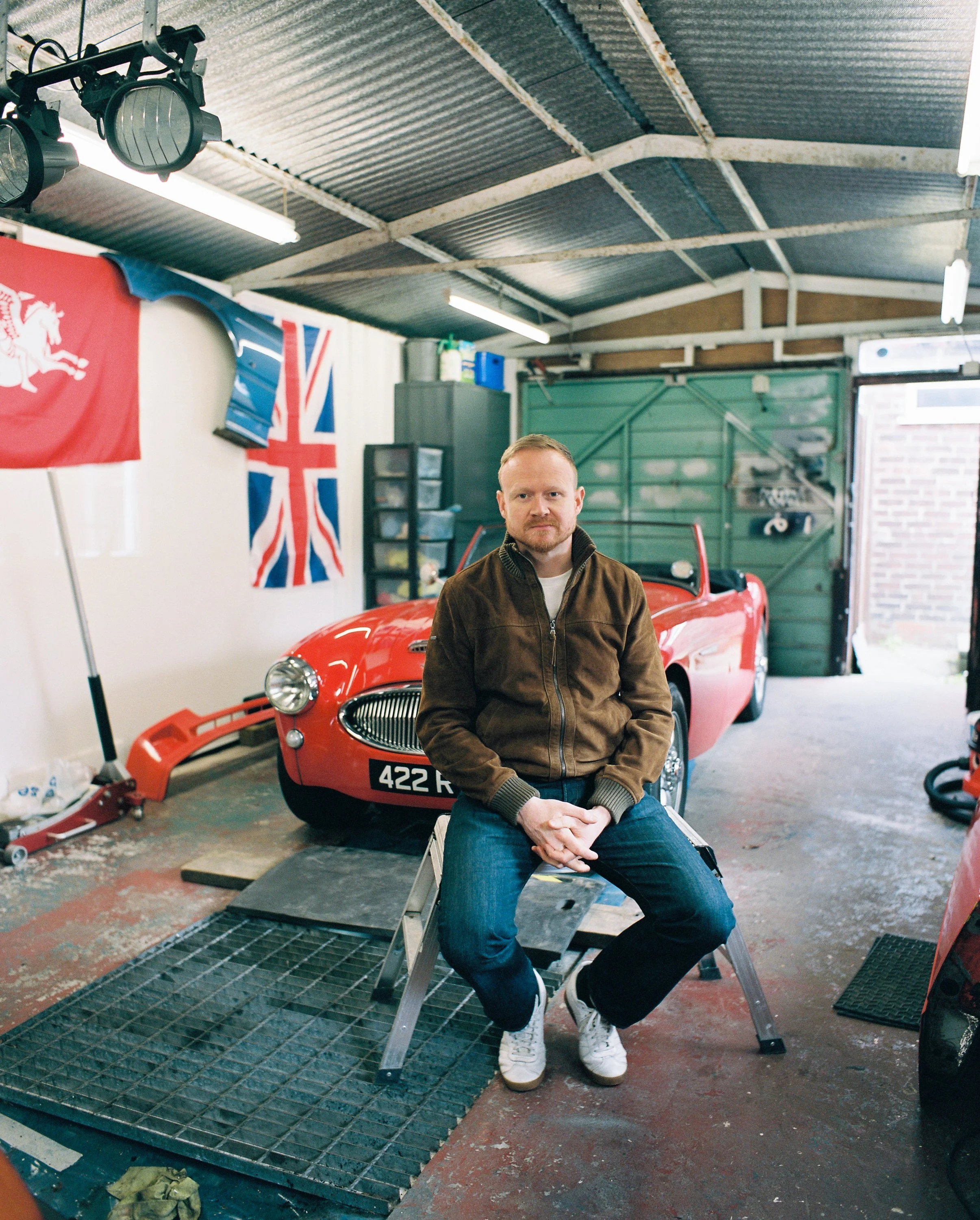 A man sitting on a small stool in a garage with a red vintage sports car in the background and flags on the wall.