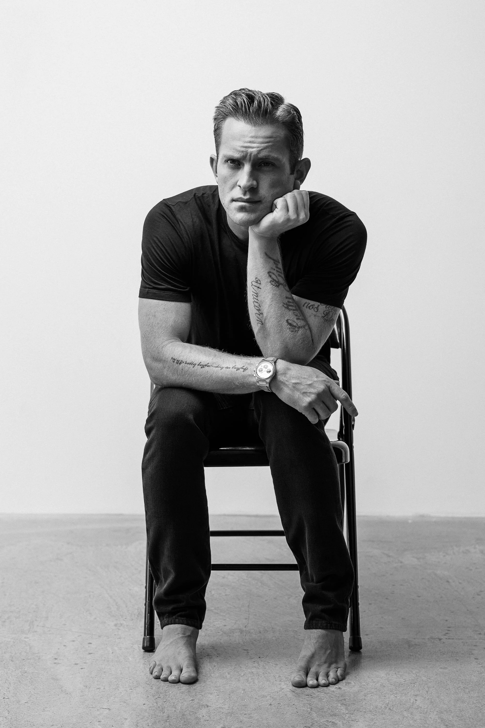 Black and white photo of a young man sitting on a chair, barefoot, wearing a black t-shirt and jeans. He rests his chin on his hand and looks into the camera with a serious expression.